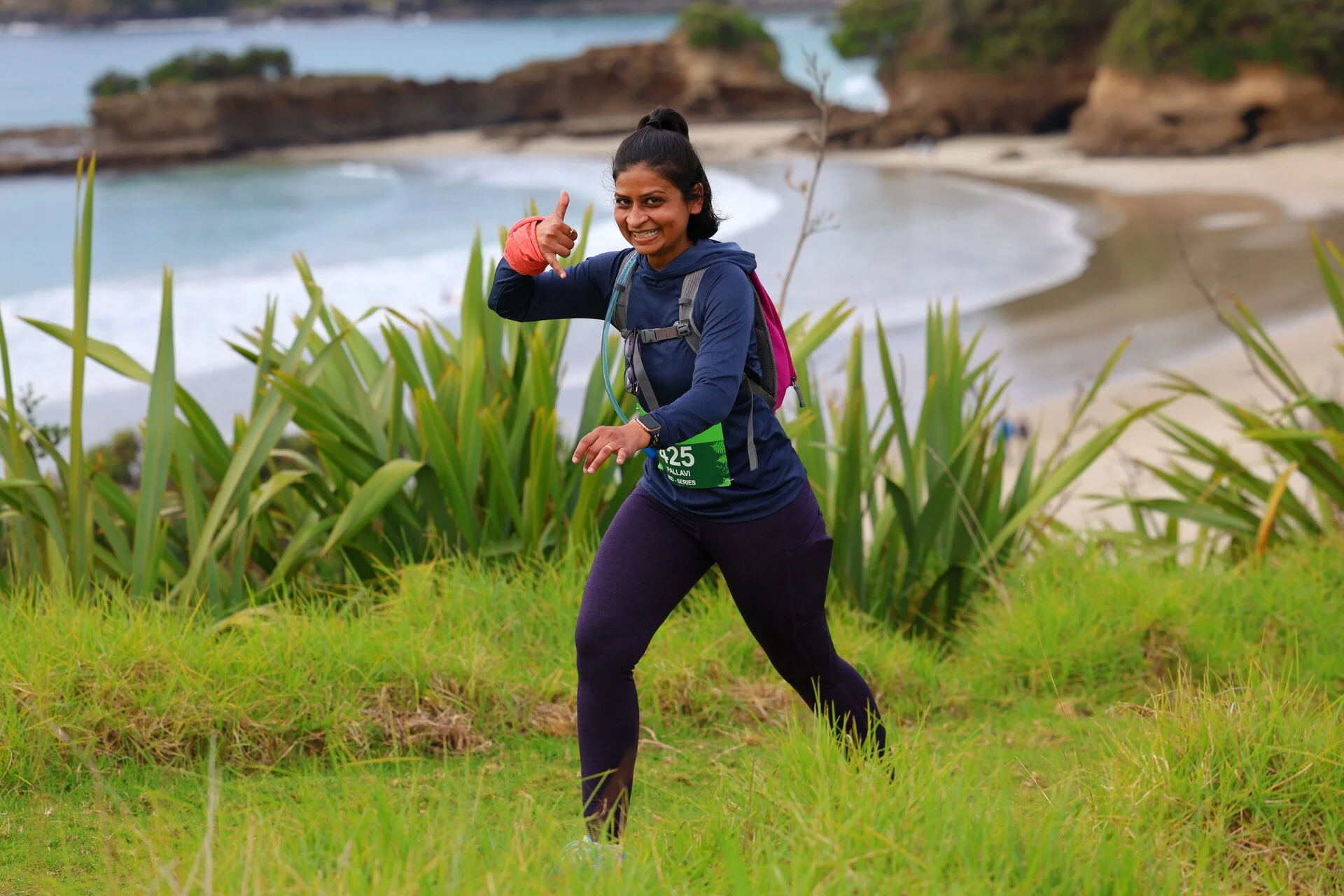 A woman in athletic gear running along a grassy path near a beach with rocky cliffs and ocean in the background, smiling and giving a thumbs-up.
