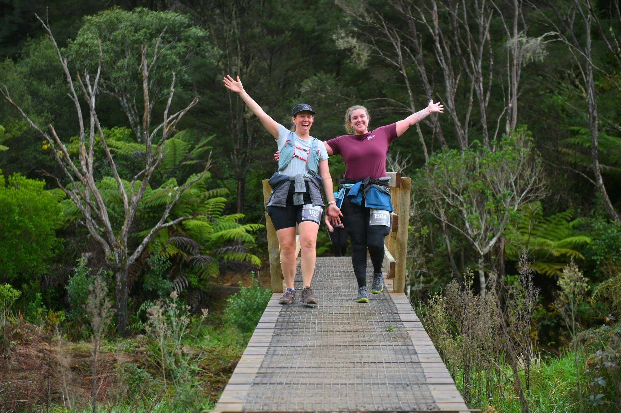 Two women joyfully walking on a wooden bridge in a lush forest, arms raised and smiling.