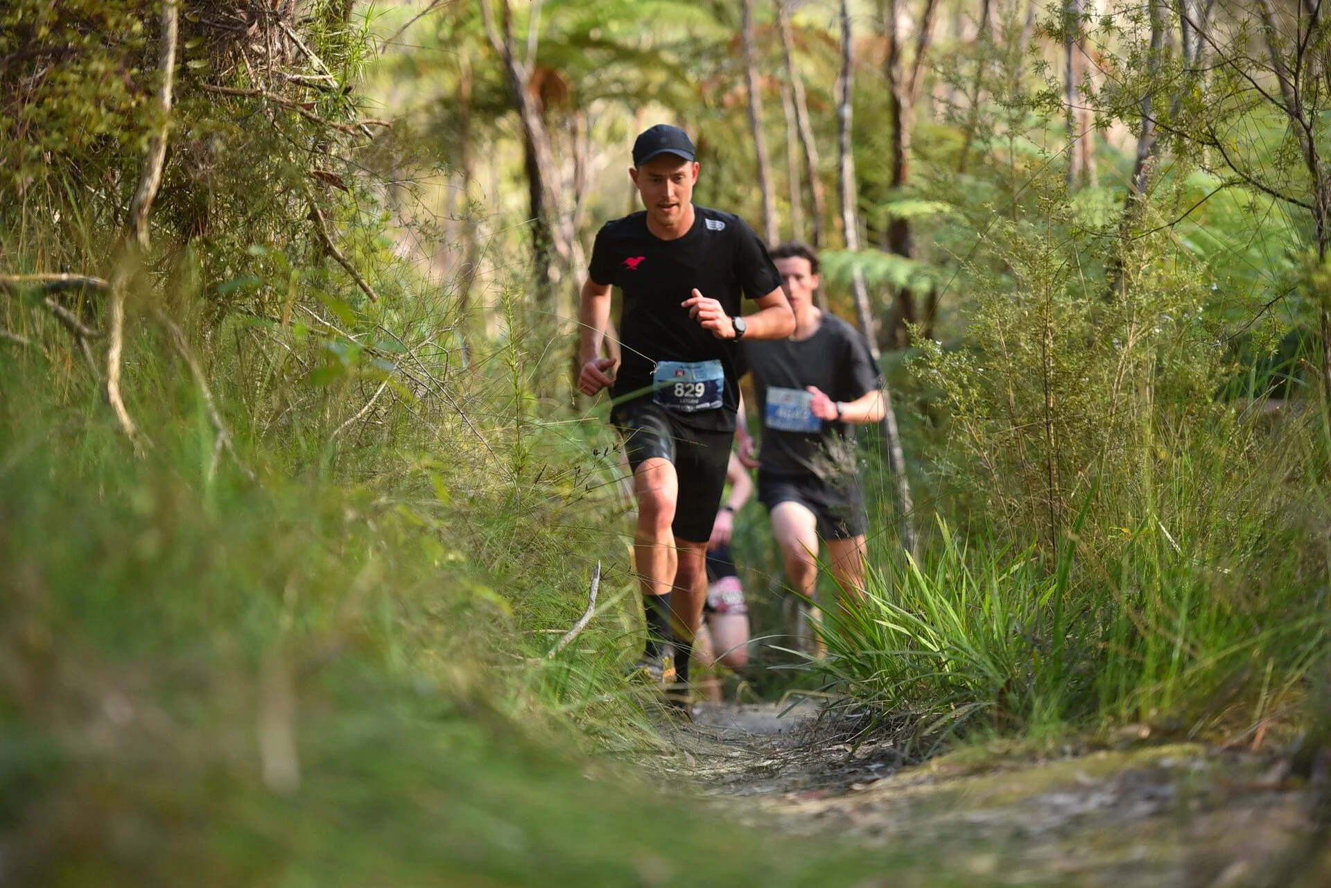 Two men running on a trail through a lush green forest, participating in a race or marathon.