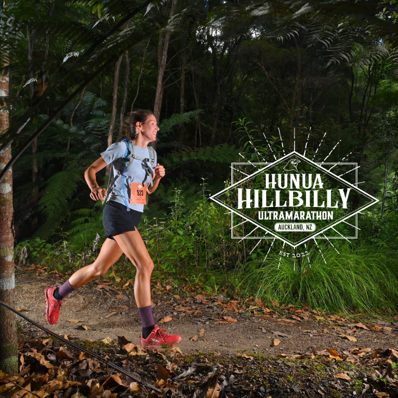 A woman running on a trail through a forest during an ultramarathon race, wearing a race bib and athletic gear, with a logo for the 'Hunua Hillbilly Ultramarathon' in Auckland, New Zealand, visible in the image.