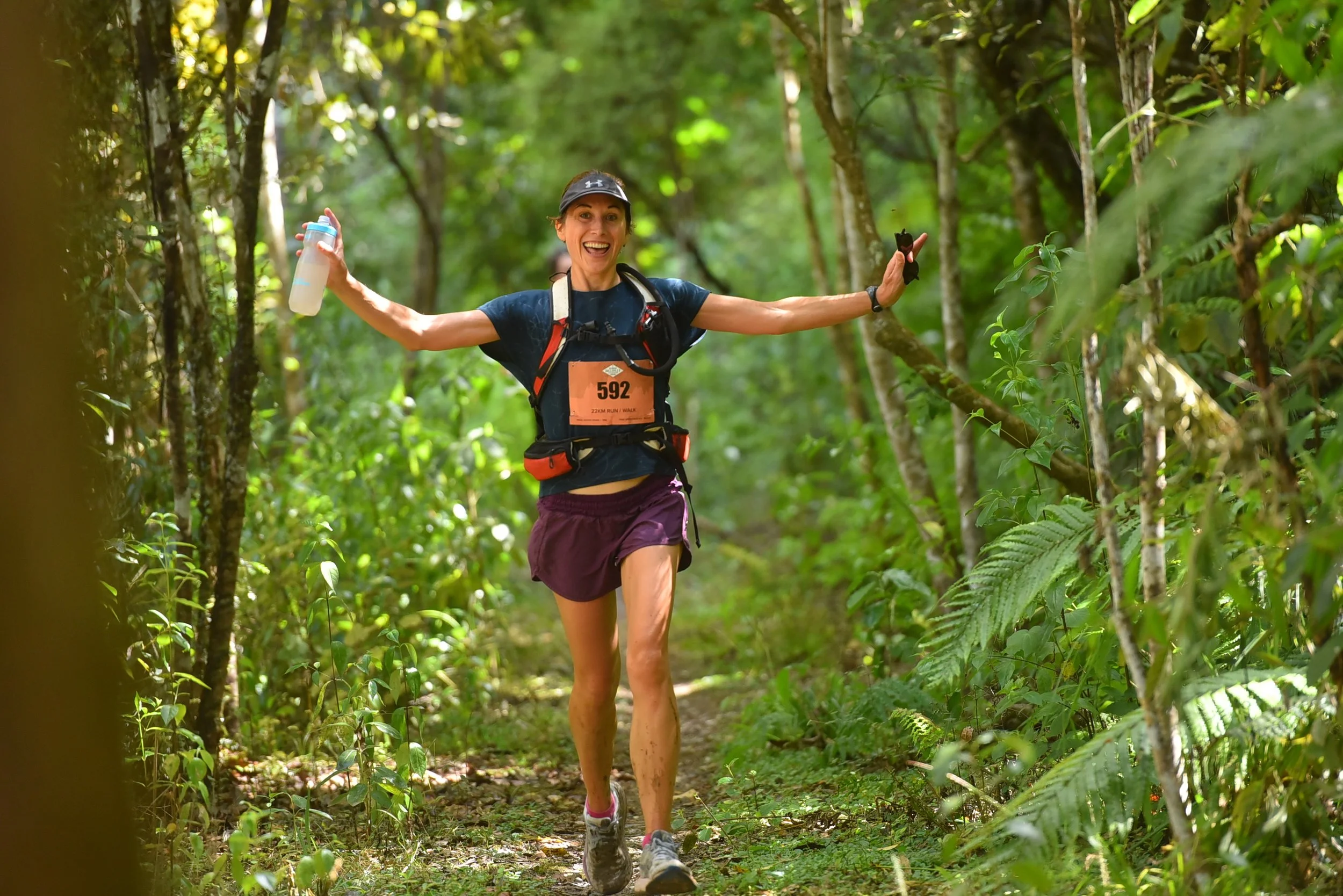 A woman running through a forest trail with arms spread open, smiling, holding a water bottle in one hand, wearing running gear, a race bib, and a hydration pack.