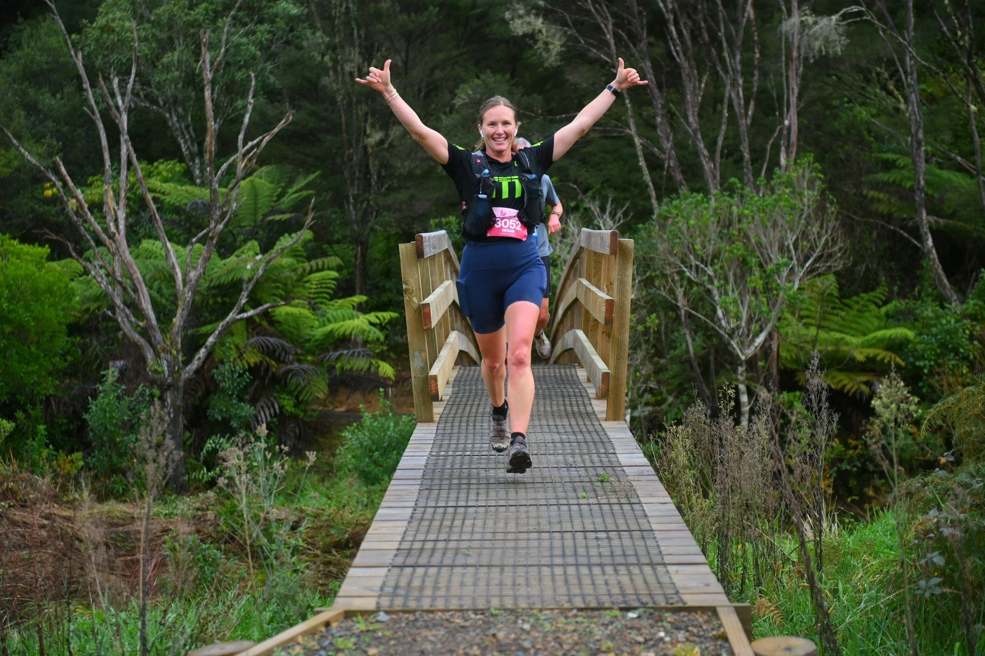 A woman joyfully running on a small wooden bridge in a lush green forest, raising her arms in a celebratory gesture.