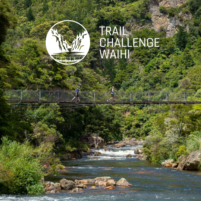 Hikers crossing a suspension bridge over a river in a lush green canyon during daytime, with a logo and text reading "Trail Challenge Waihi" overlayed.