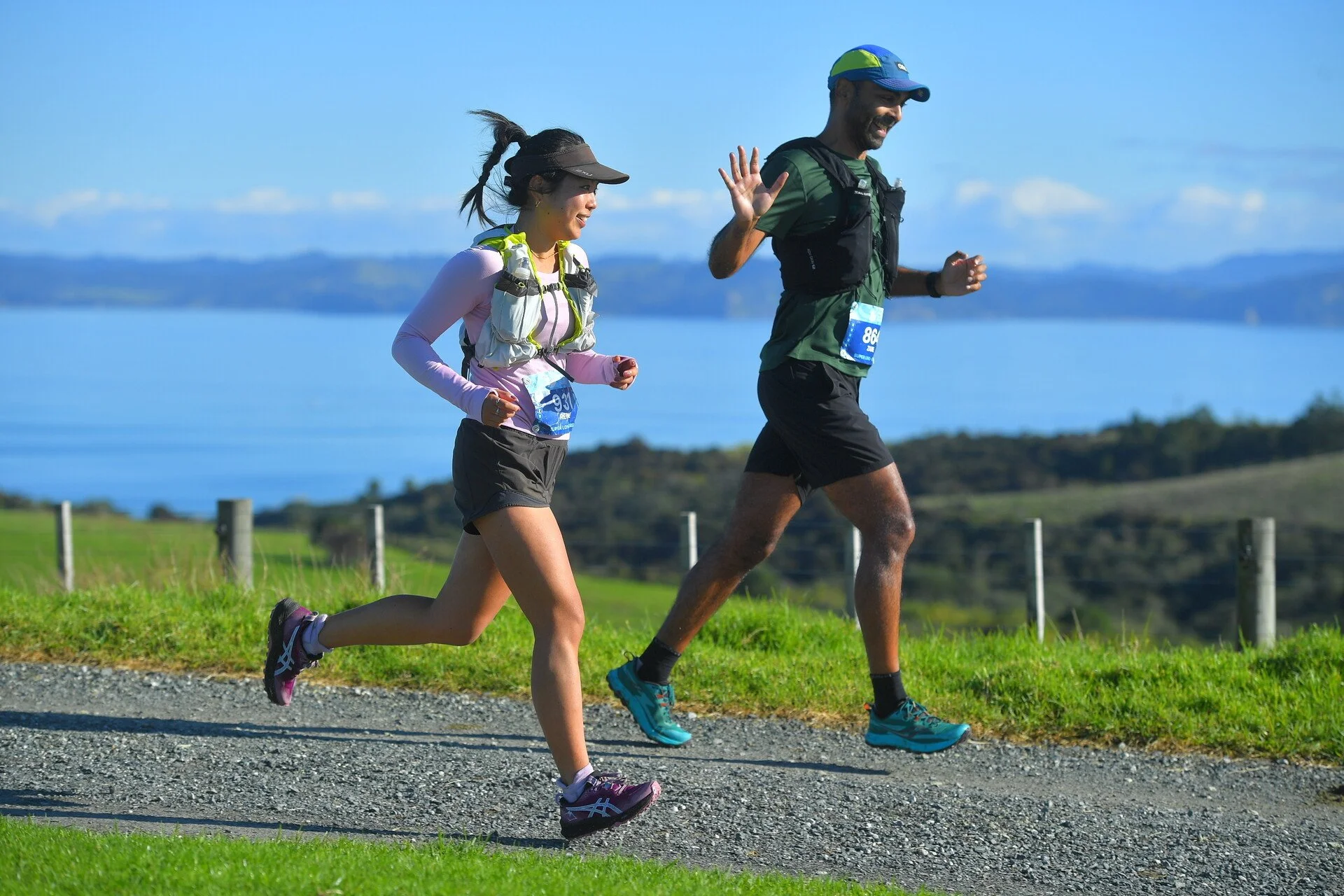 Two runners, a woman and a man, running outdoors on a gravel trail with scenic water and hills in the background.