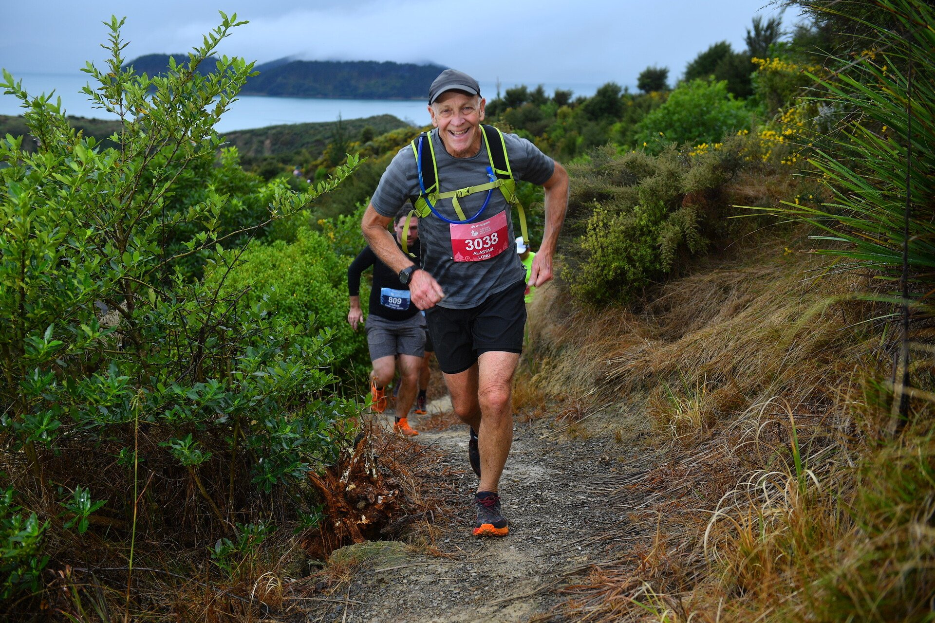 An older man running on a trail through greenery near a body of water, smiling with a race bib number 3038 on his chest, wearing a gray cap, black shorts, and a hydration pack.