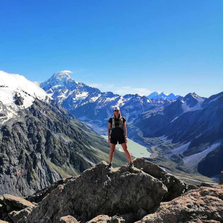 A woman standing on a large rock in a mountain landscape with snow-capped peaks and a clear blue sky.