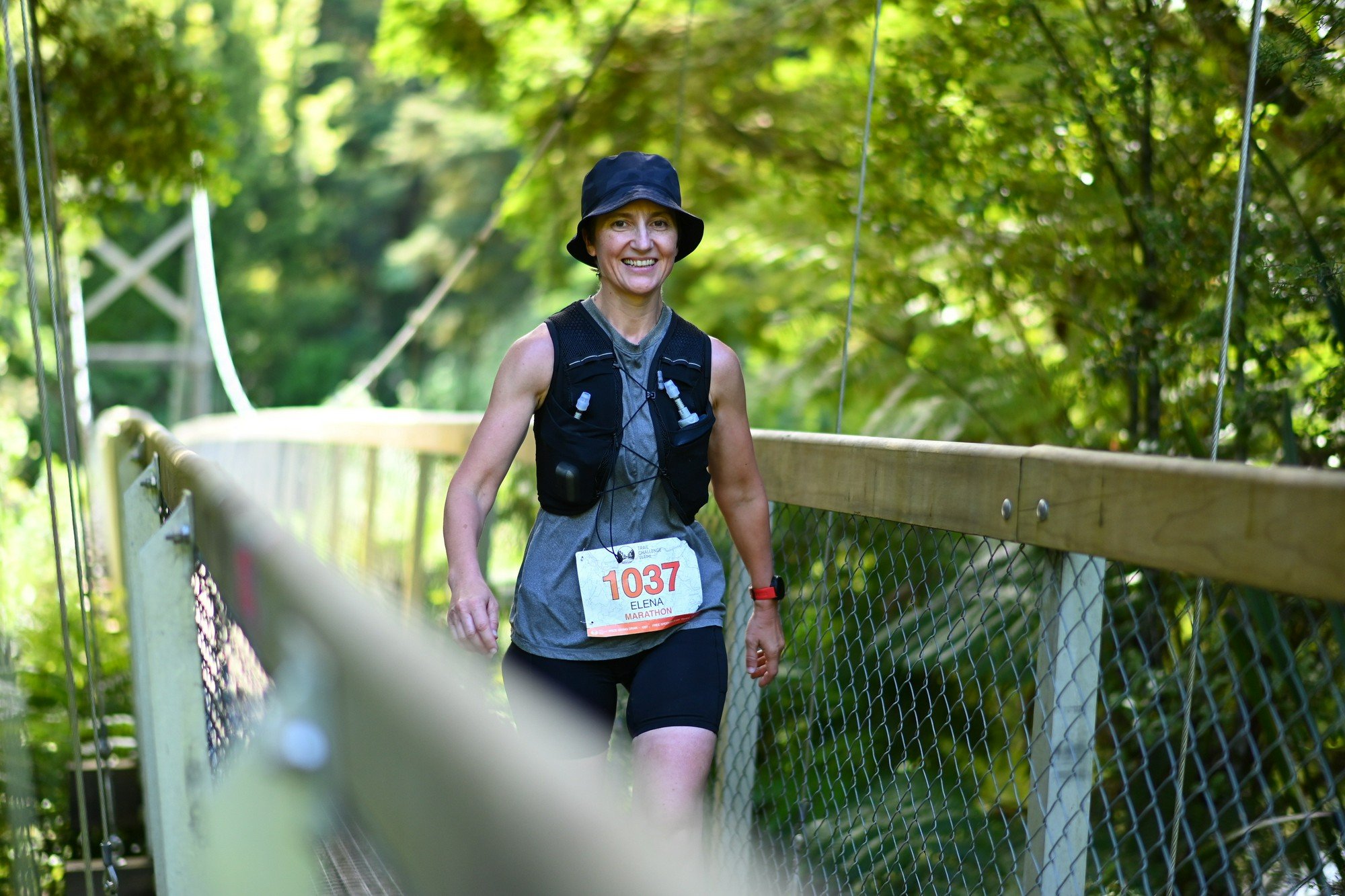 A woman smiling while running outdoors on a trail, wearing a hat, a race bib with the number 1037, a hydration vest, and black shorts, surrounded by greenery.