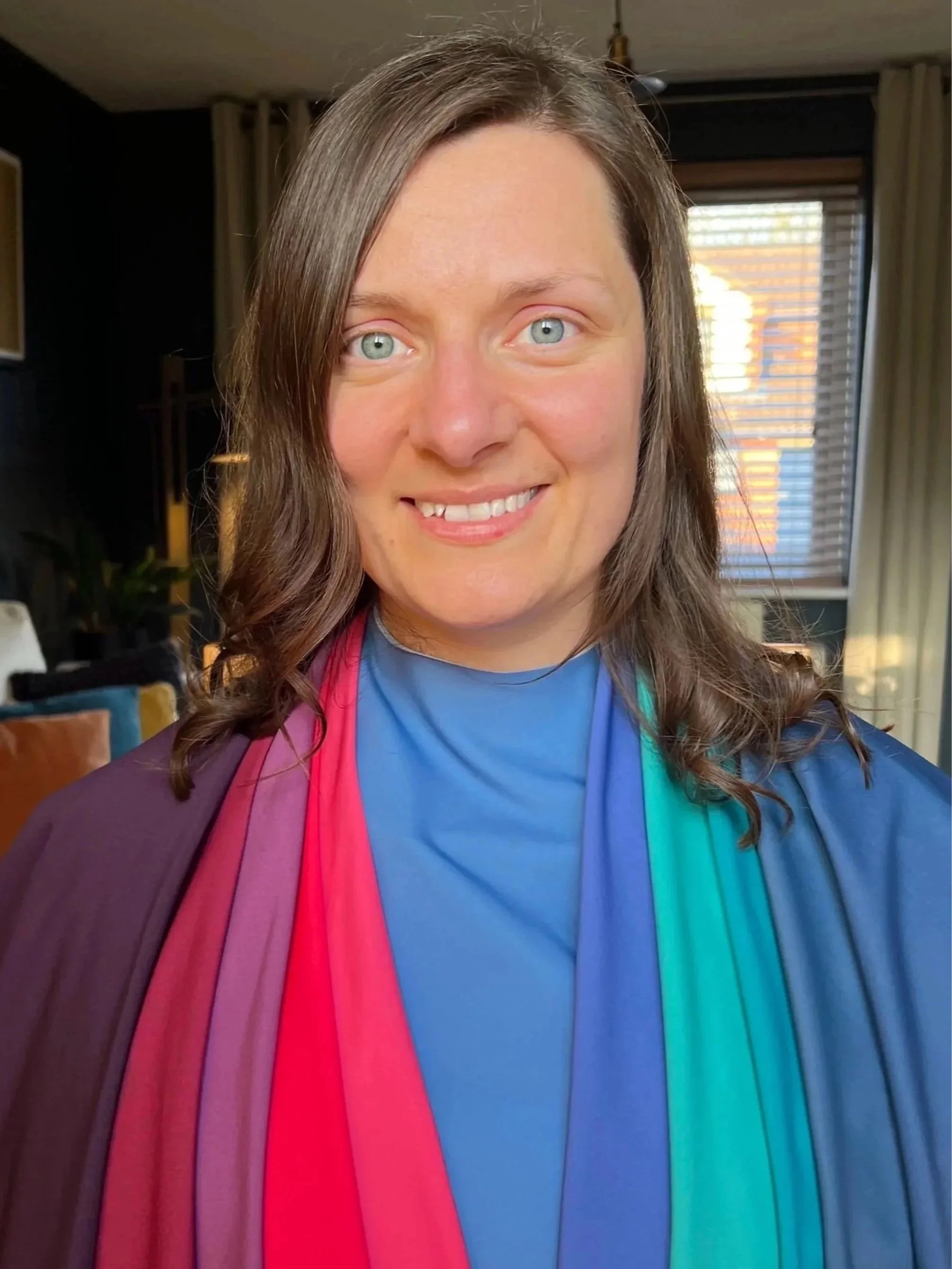 A woman with shoulder-length brown hair and blue eyes smiling at the camera. She is wearing a blue shirt and draped in a rainbow-colored graduate stole. The background shows a living room with curtains and window blinds.