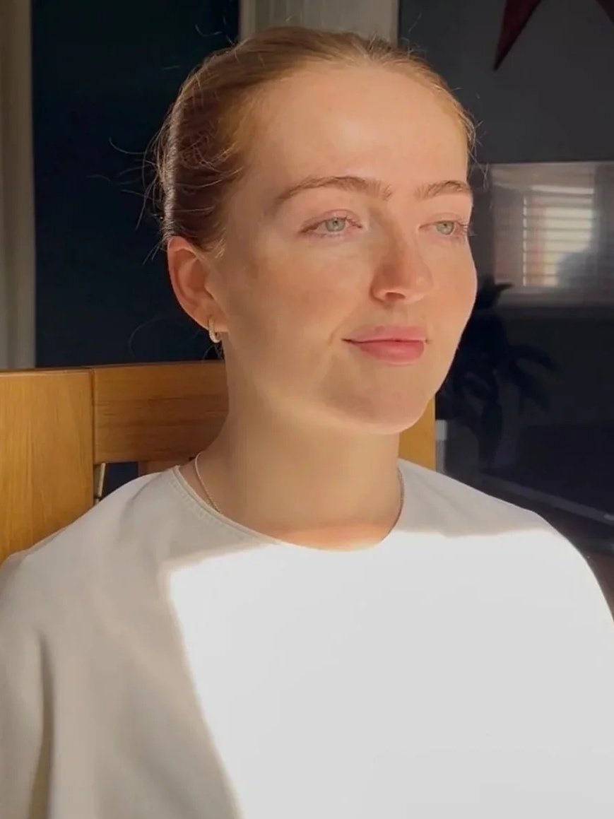 A woman with red hair pulled back, light skin, and blue eyes is sitting indoors in a chair, with sunlight shining on her face and shoulder.