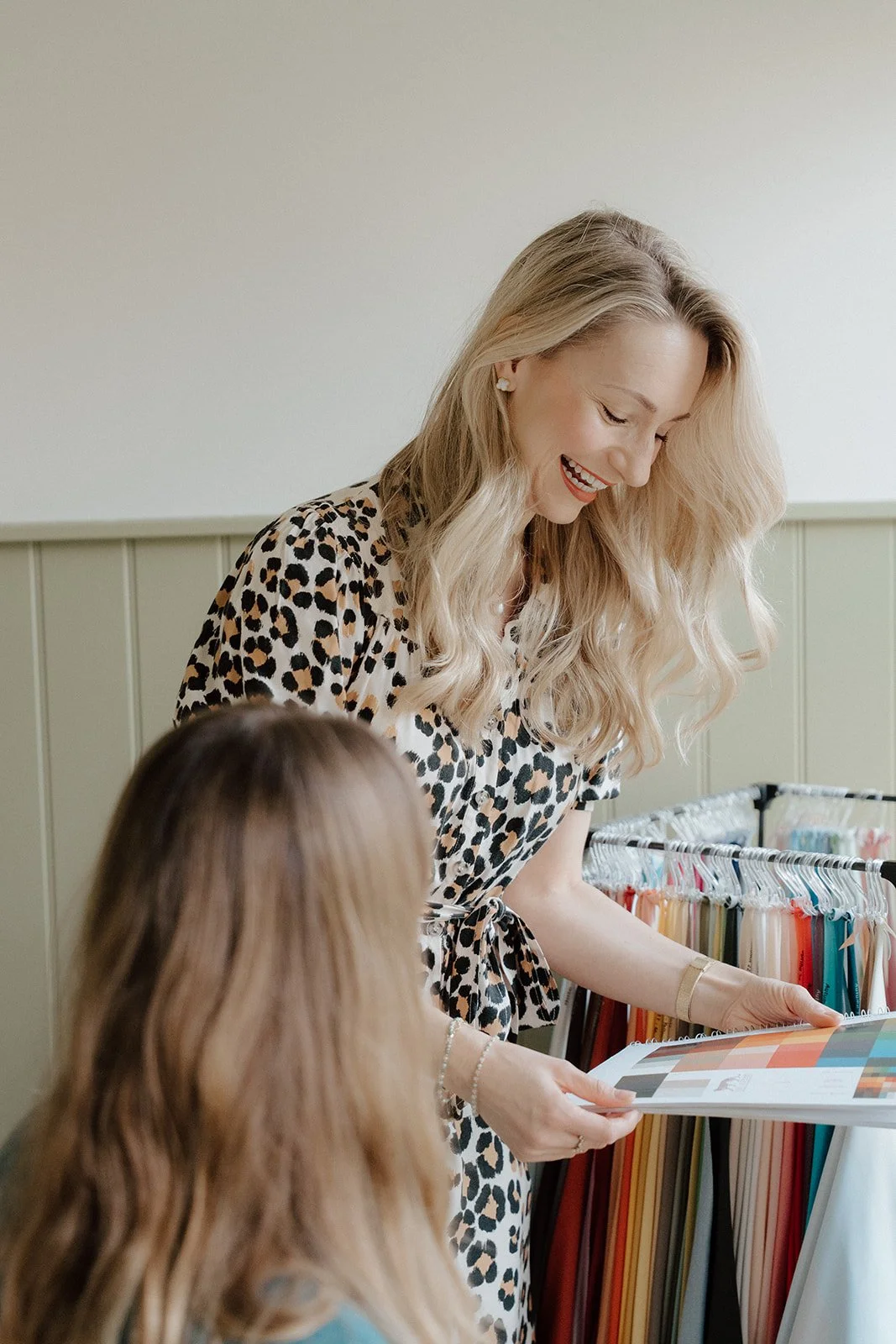 A woman with lapel-length blonde hair wearing a leopard print dress smiling and looking at a color palette held by a young girl with long hair.