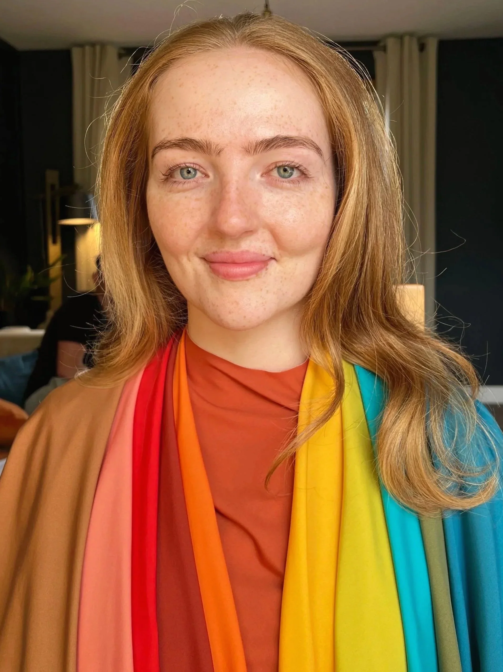 Close-up of a woman with red hair and blue eyes, smiling, wearing a multicolored rainbow scarf and a reddish-brown top, inside a warmly lit room.
