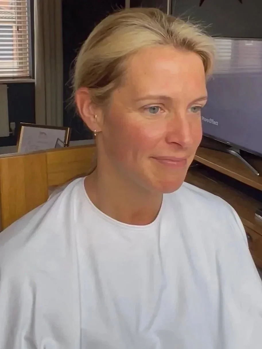 Woman with blond hair and blue eyes wearing a white shirt, sitting indoors with a television and window in the background.