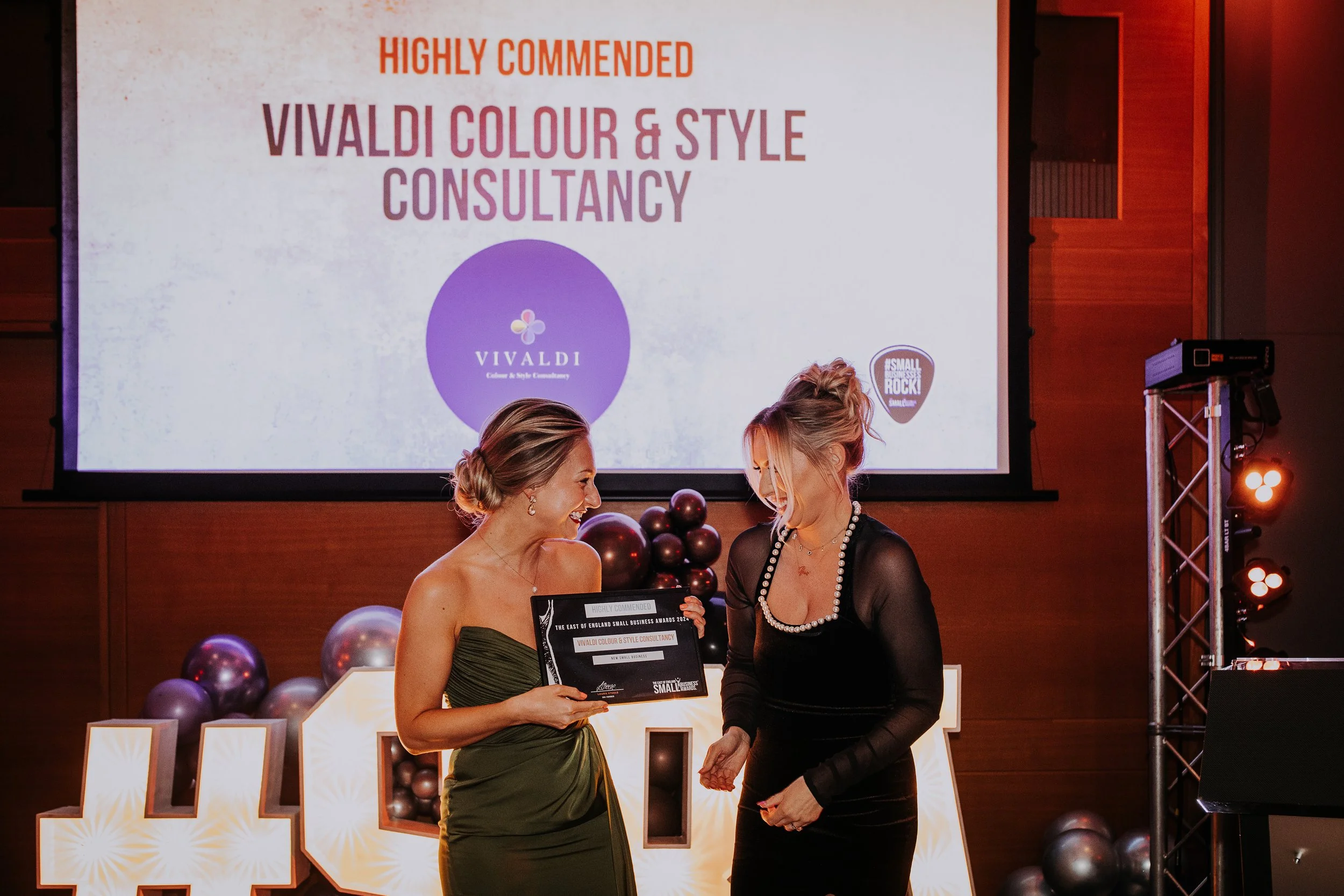 Two women are on stage at an awards ceremony, smiling and holding an award plaque. Behind them is a large screen displaying "Highly Commended Vivaldi Colour & Style Consultancy." The stage is decorated with black balloons and illuminated letters.