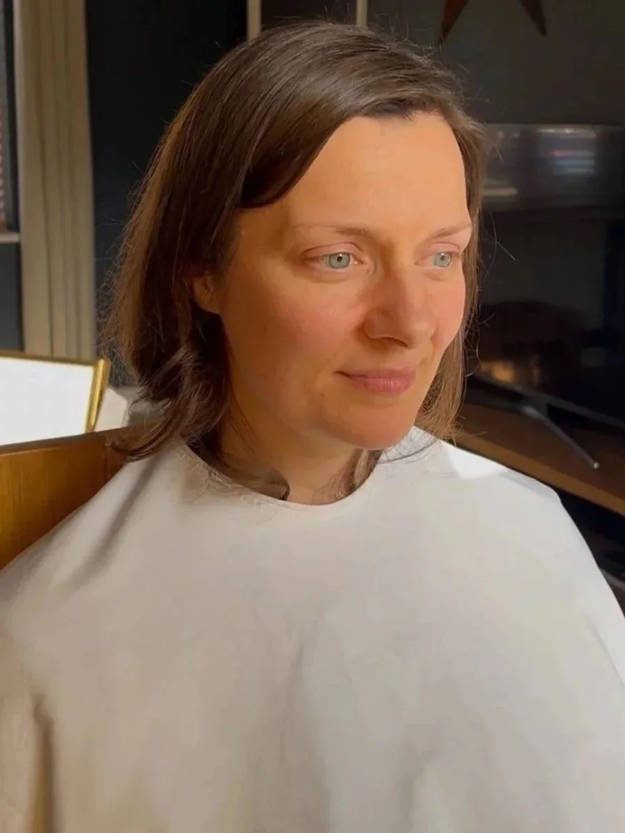 A woman with shoulder-length brown hair and blue eyes sitting in a room, wearing a light-colored shirt.
