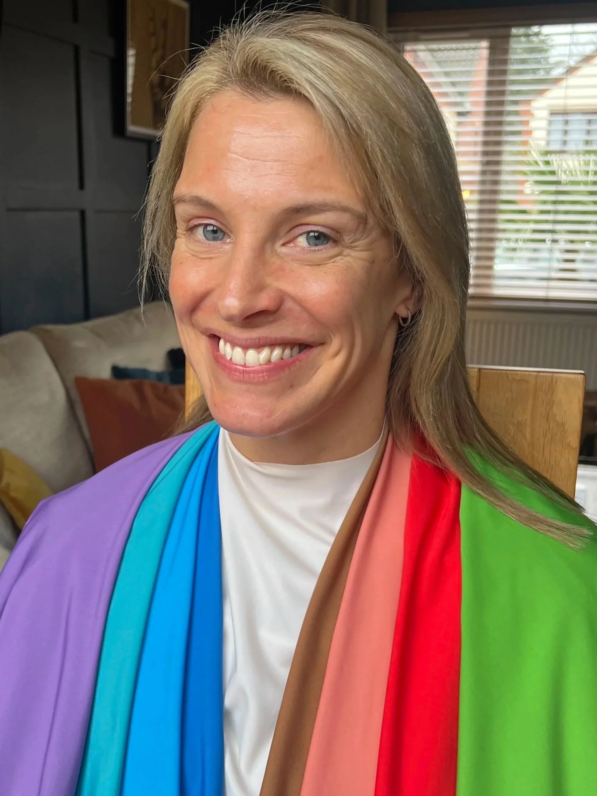 Woman smiling, seated indoors with a rainbow-colored scarf. Behind her are window blinds and a living room setting.