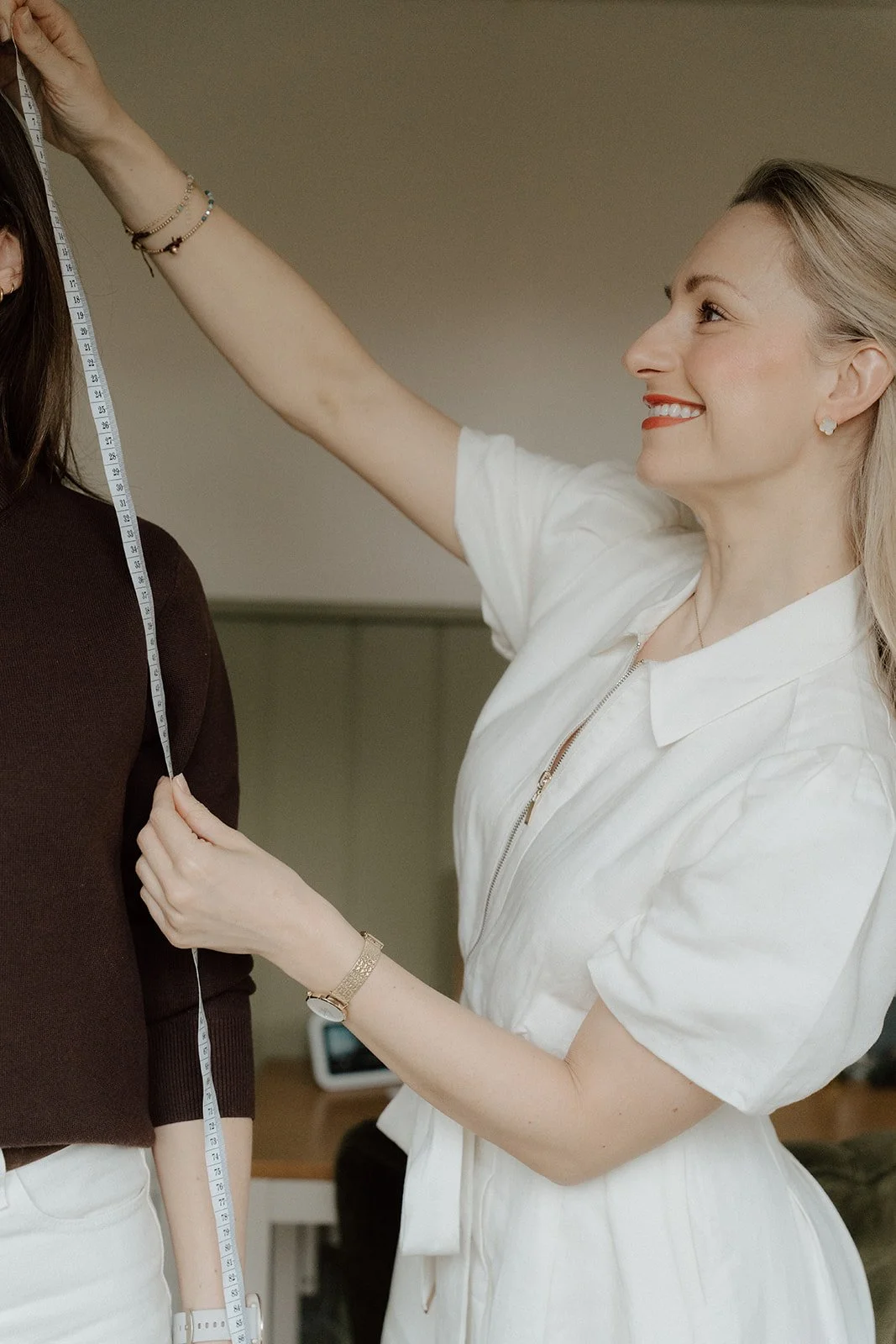 A woman in a white dress measuring the height of another woman in a dark brown sweater using a measuring tape.