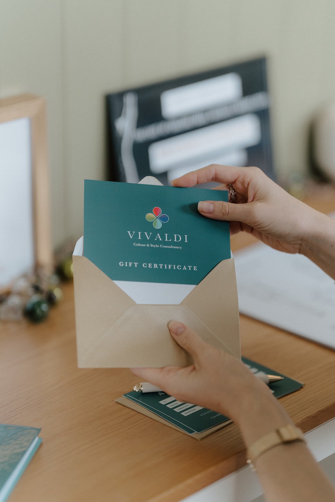 Person holding a gift certificate from Vivaldi Colour & Style Consultancy, inside a beige envelope, at a wooden desk with some stationery items and blurred background.