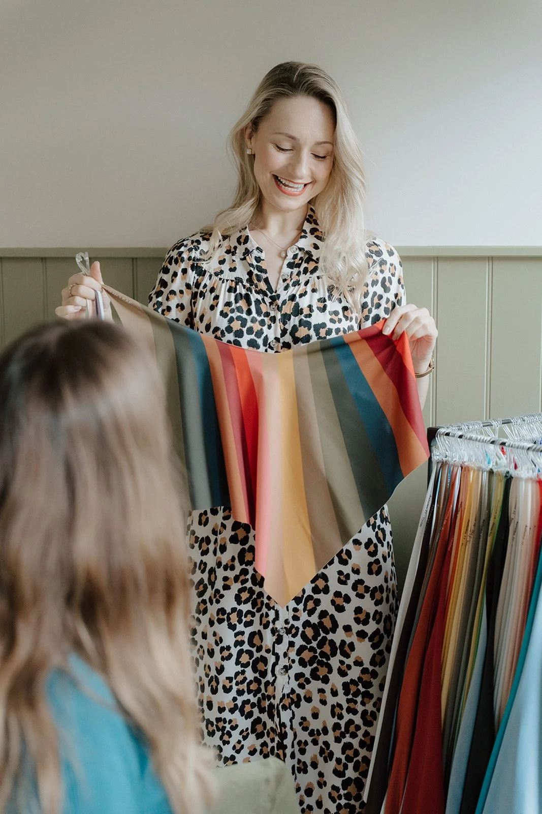 A woman in a leopard print dress holding a rainbow-colored fabric, showing it to a person with blonde hair inside a fabric store.