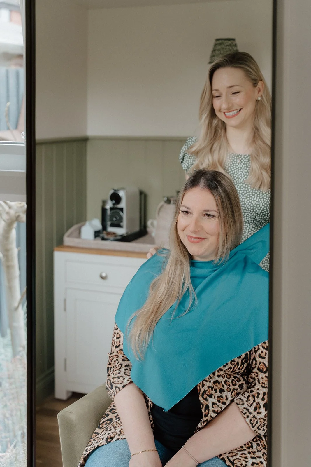 Two women, one seated and one standing behind her, looking into a mirror. The seated woman has long blonde hair, a leopard print top, and a blue cape. The standing woman has shoulder-length blonde hair and is smiling, wearing a green patterned top.