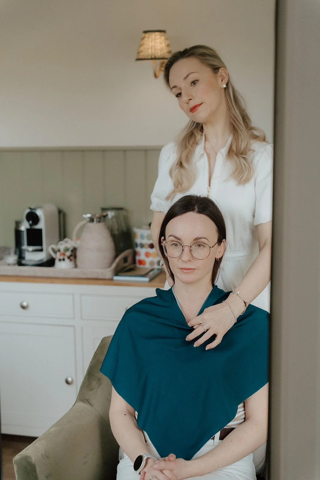 A woman with dark hair wearing glasses and a blue shawl is sitting in a chair, while another woman with blonde hair in a white shirt stands behind her touching her shoulder.