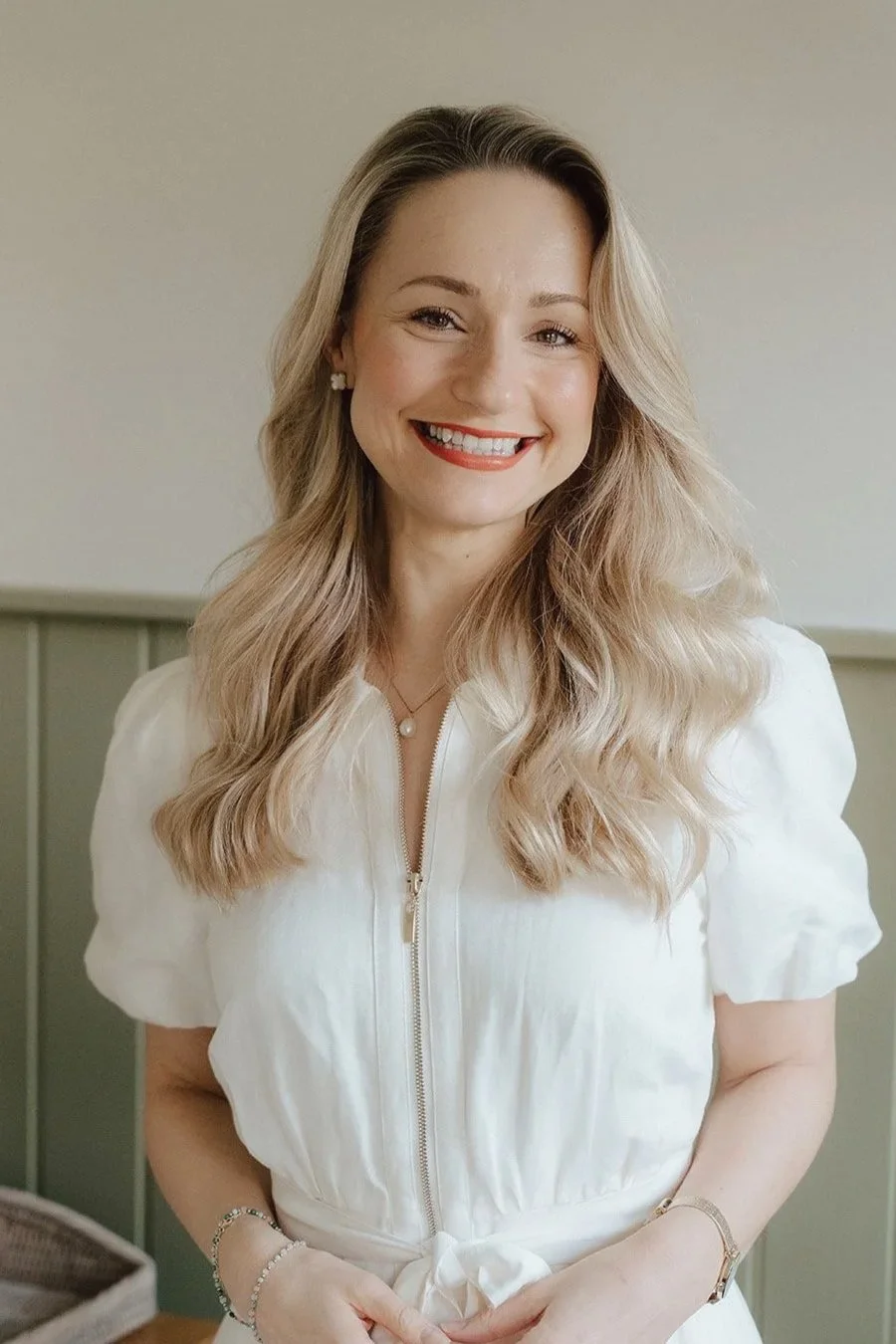 A smiling woman with wavy blonde hair, wearing a white zip-up dress and pearl jewelry, standing indoors in front of a light-colored wall.
