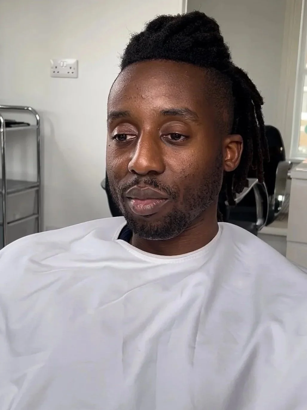 A man with dreadlocks and a beard sitting in a chair at a clinic or salon, wearing a white gown.