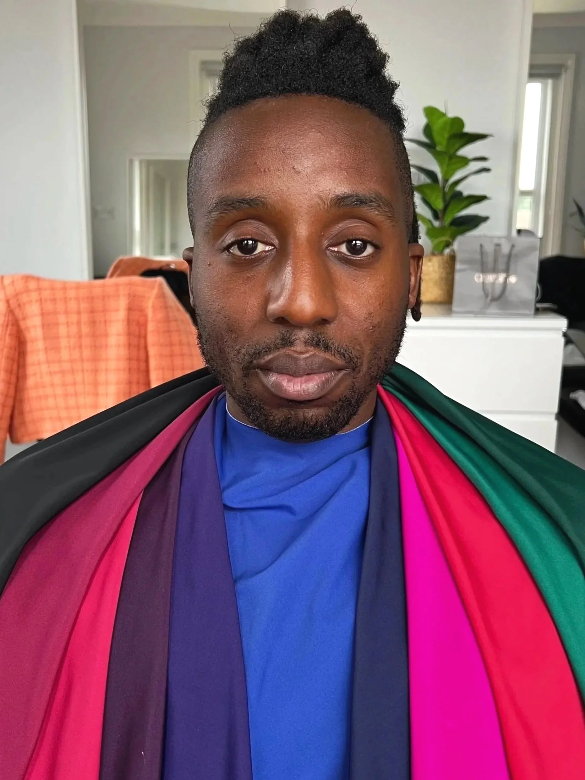 A person with short curly hair and a beard, wearing a rainbow pride flag draped over their shoulders, standing in a room with a white dresser, potted plant, and mirror in the background.