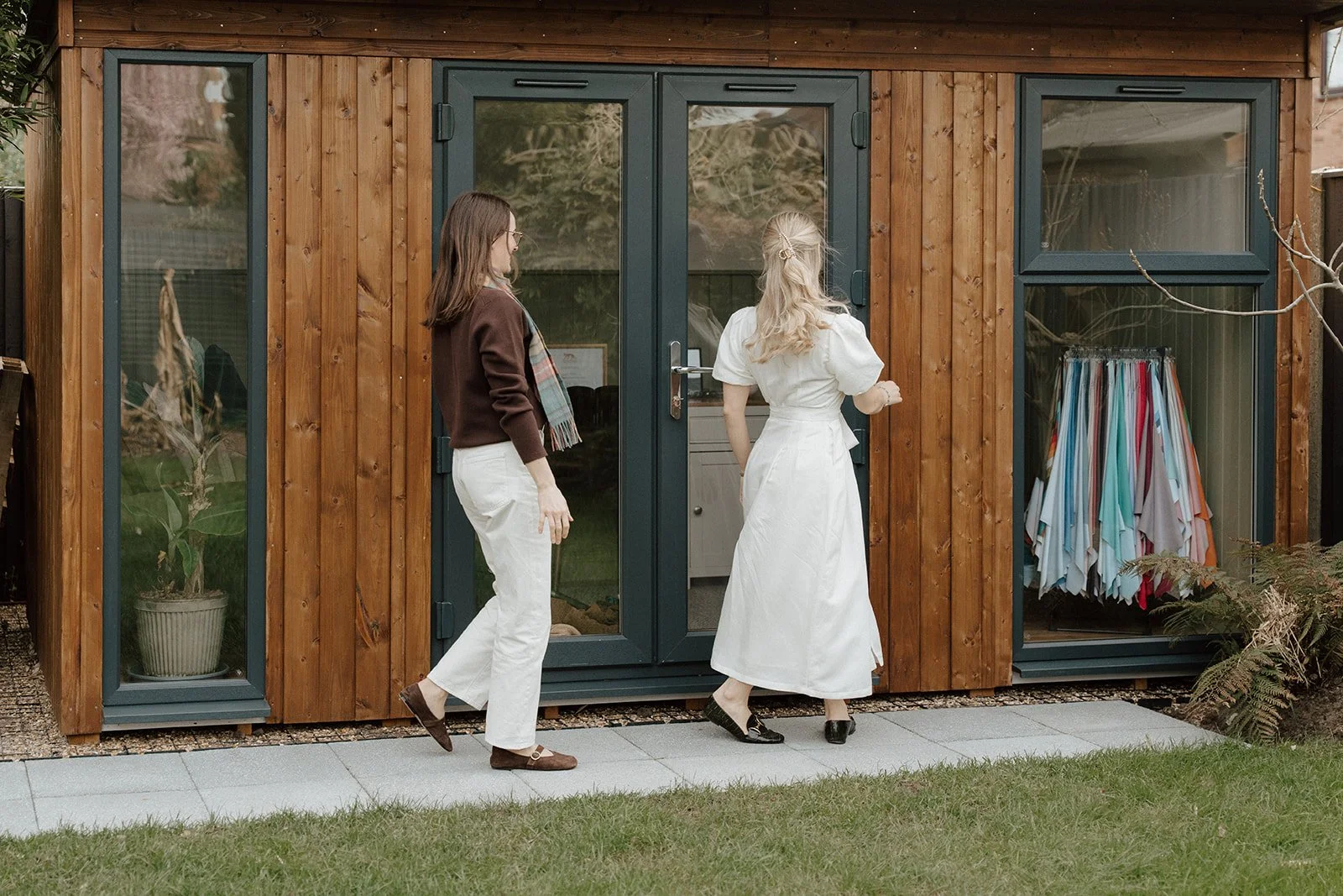 Two women, one with long brown hair wearing a brown sweater and white pants, and the other with long blonde hair in a white dress, standing outside a wooden shed with glass doors and windows, engaging in conversation.