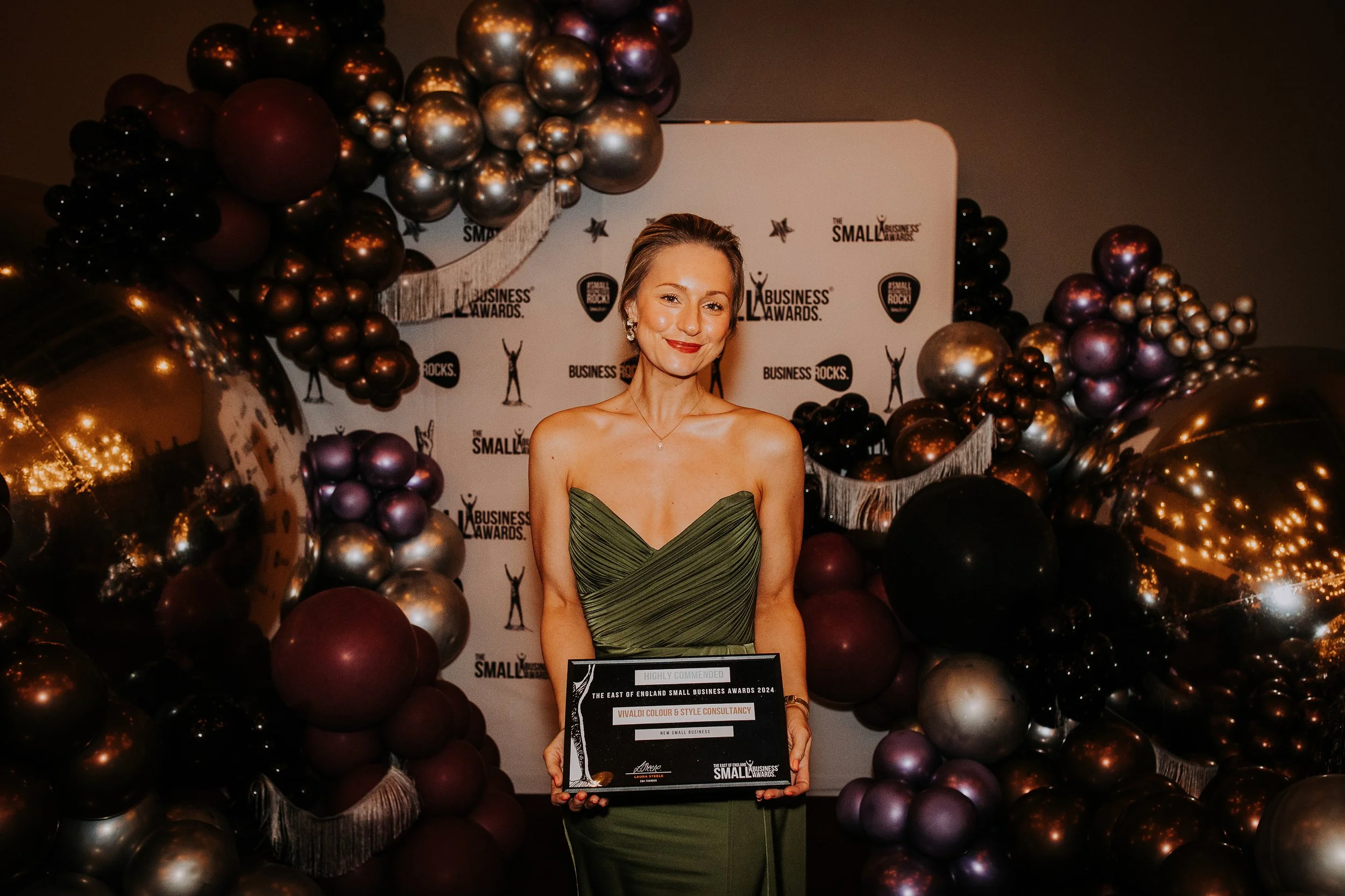 Woman in an elegant green dress holding an award, standing in front of a decorated backdrop with balloons and logos at a small business awards event.