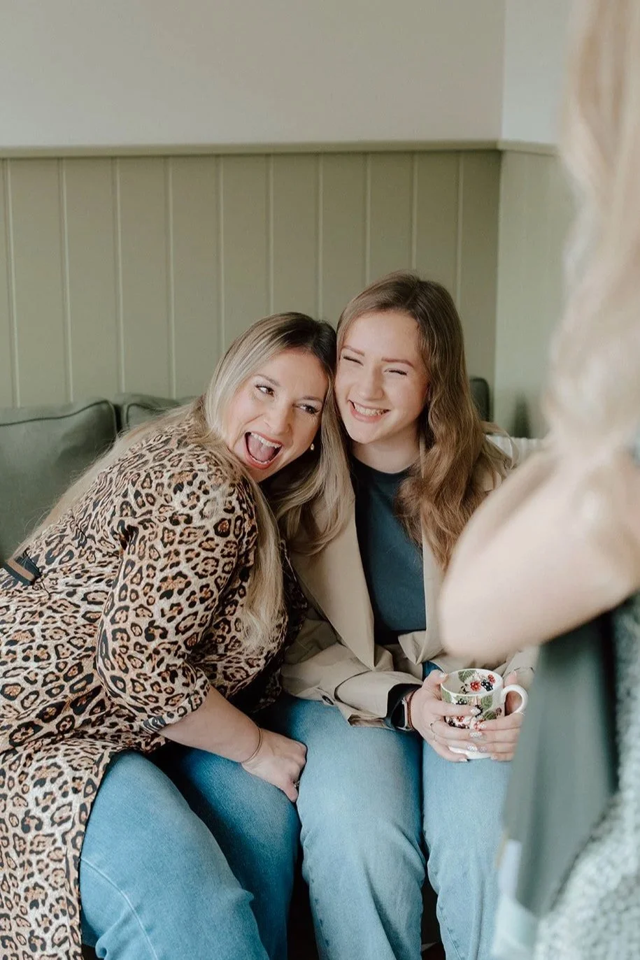Two women sitting on a green couch, smiling and laughing, with one holding a floral mug. One woman is wearing a leopard print top, and the other is wearing a beige blazer and a dark top.