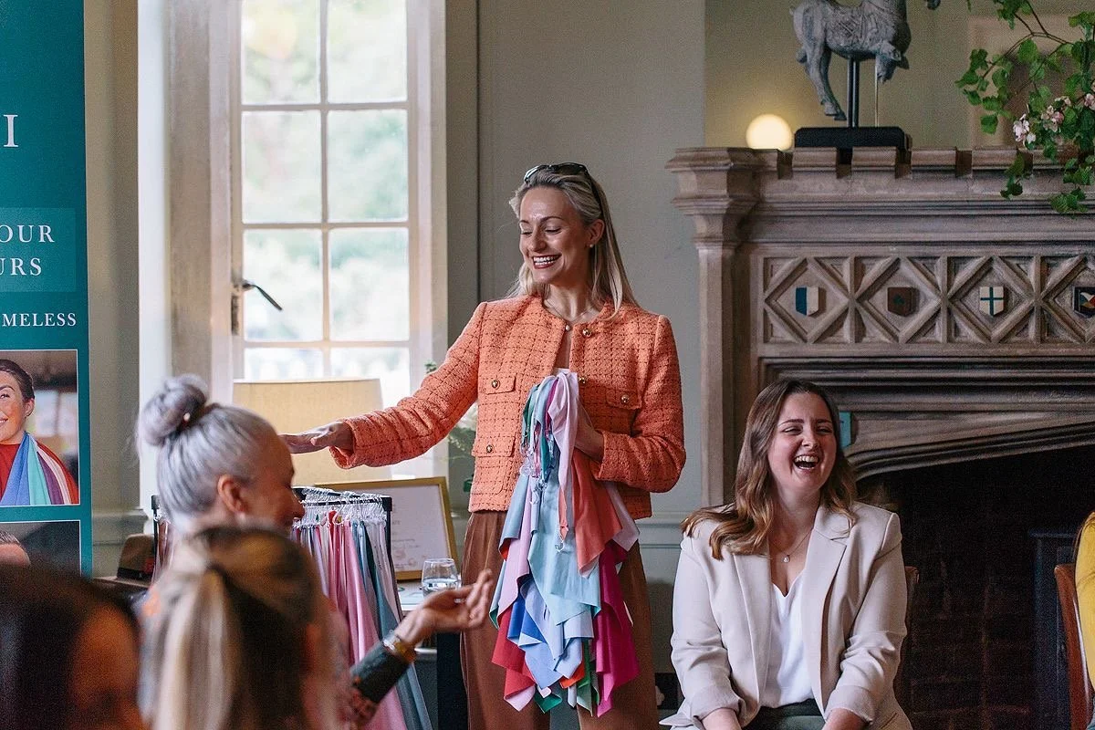 A woman smiling and holding a color swatch book at a clothing swatch display during a fashion or fabric event, with other people sitting and engaging around her.