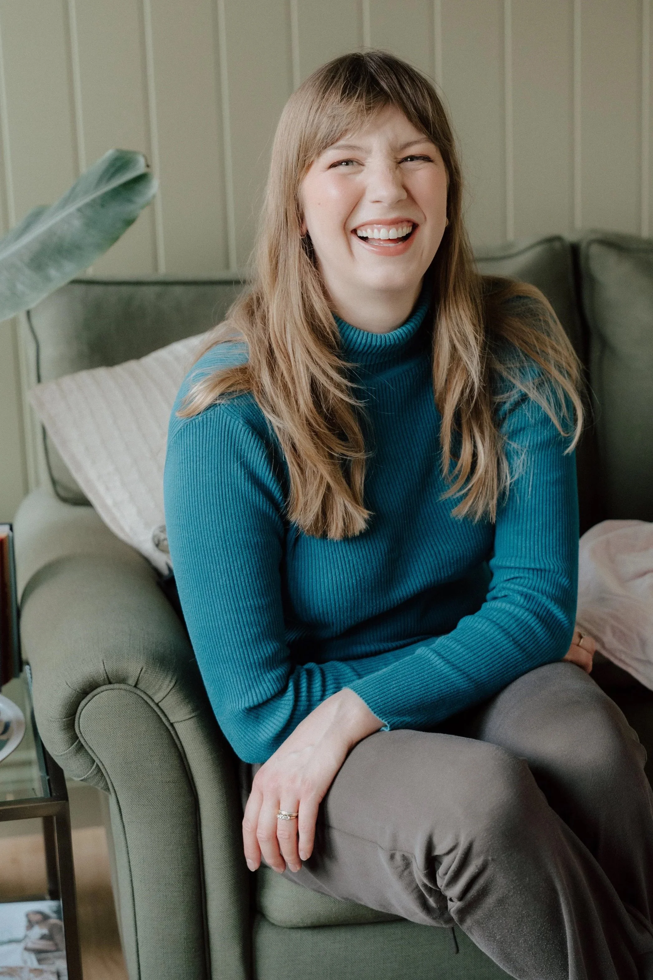 A woman with light brown hair smiling while sitting on a green sofa in a cozy living room.