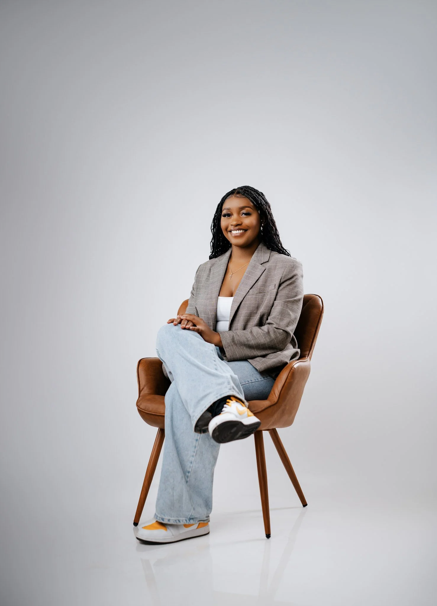 A woman sitting on a brown chair against a plain gray background, smiling, wearing a gray blazer, white top, light blue jeans, and sneakers.