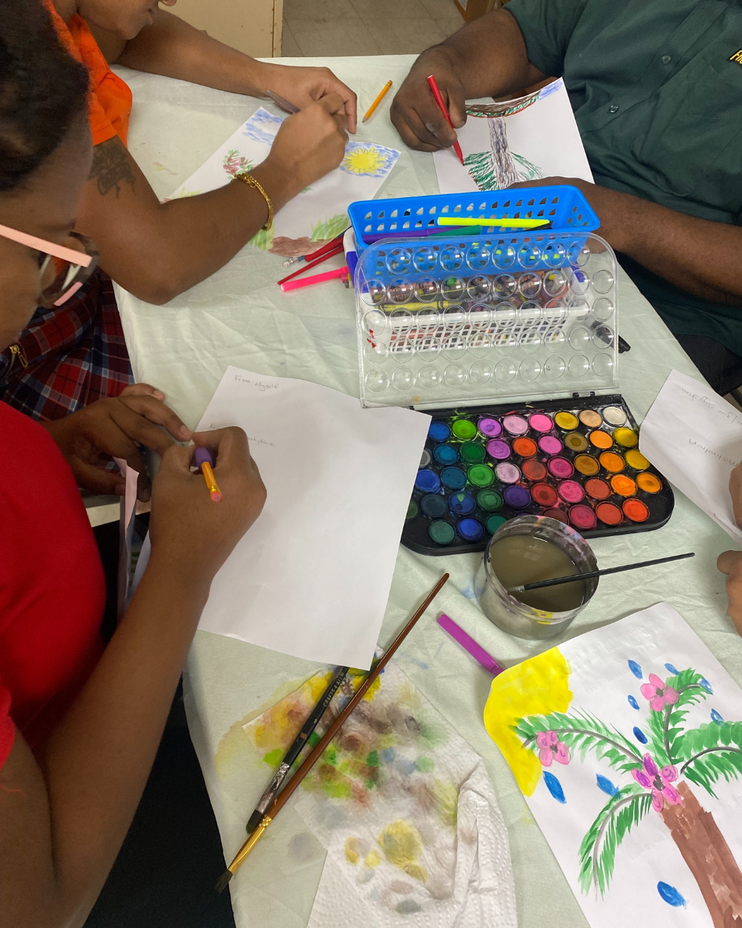 Children and adults painting and drawing with watercolors, paintbrushes, and paper, featuring a painted palm tree with pink flowers, a yellow sun, and blue raindrops on one paper.