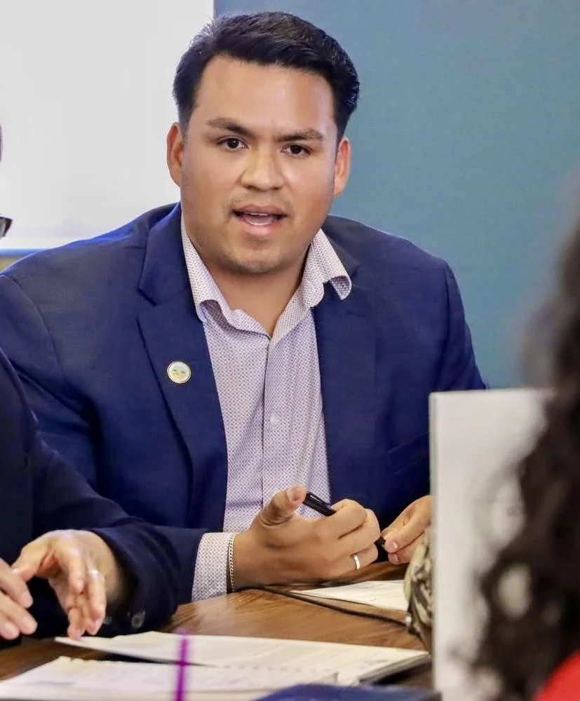 A man in a suit and dress shirt speaking during a meeting, with papers and a pen on the table.