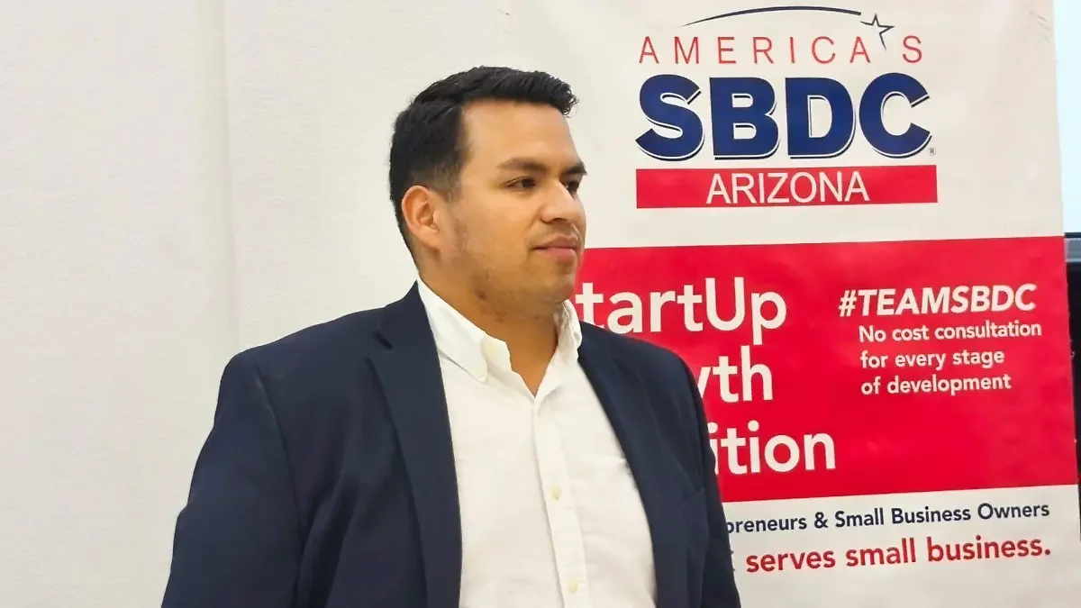 A man in a white shirt and dark blazer standing in front of a red and white banner that reads 'America's SBDC Arizona', with hashtags and descriptions about startup growth and small business support.