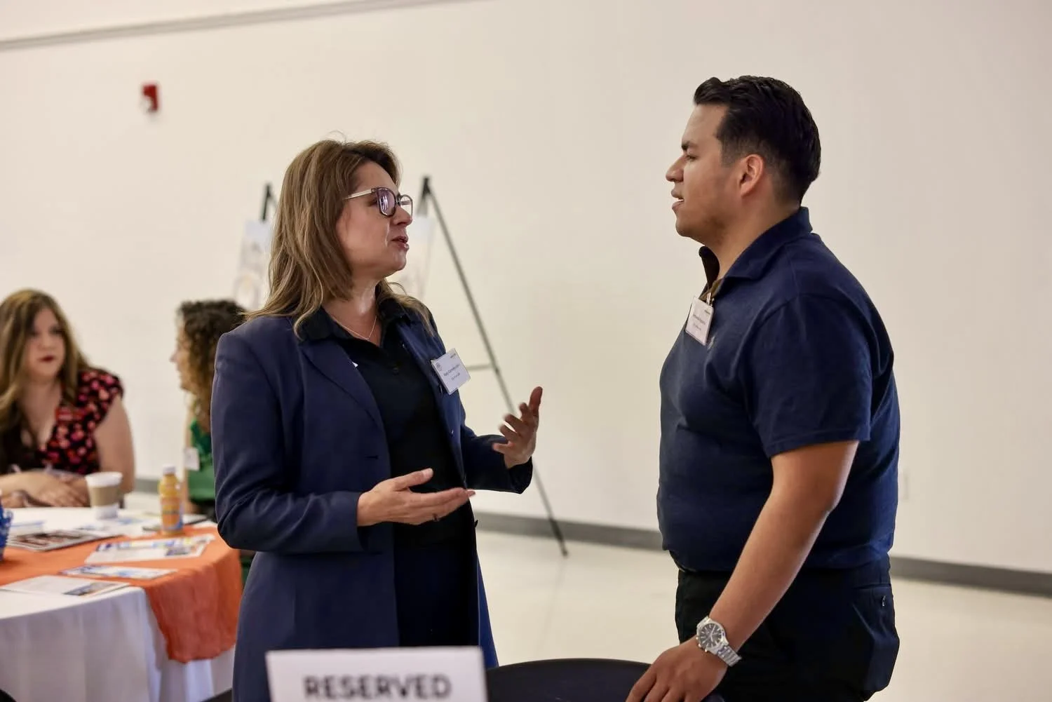 Two people, a woman with glasses and a man, are having a conversation at a professional event. The woman is wearing a dark blazer and the man is wearing a dark shirt with a name tag. In the background, a table displays brochures and there is a 'RESER