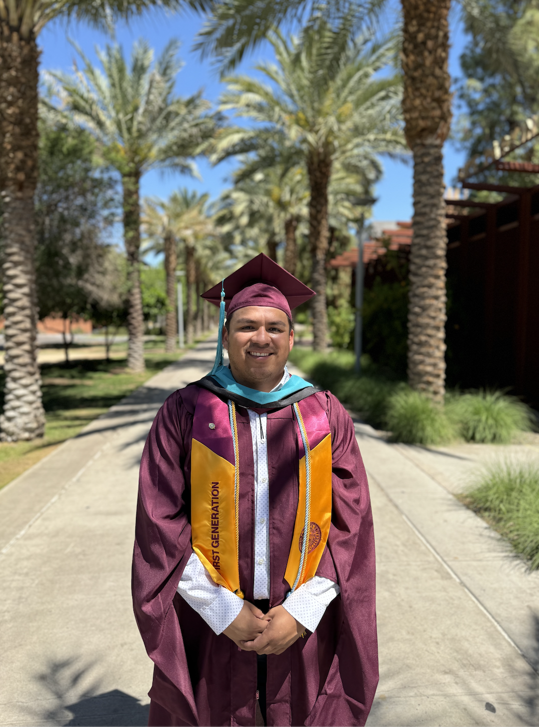 A young man in a maroon graduation gown and cap standing on a sidewalk lined with tall palm trees, smiling at the camera.