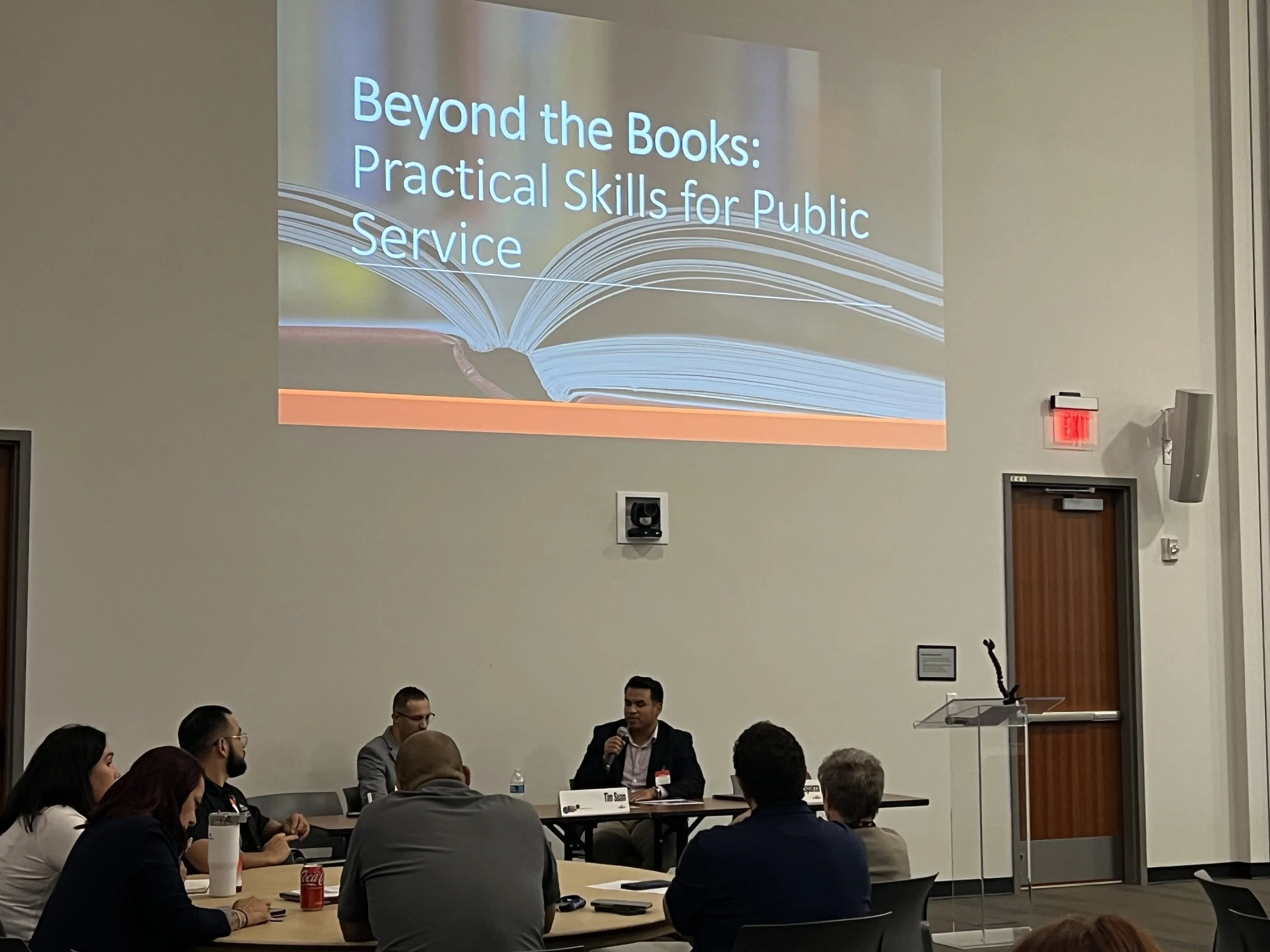 A presentation in a conference room titled "Beyond the Books: Practical Skills for Public Service" with a speaker sitting at a table and audience members listening.