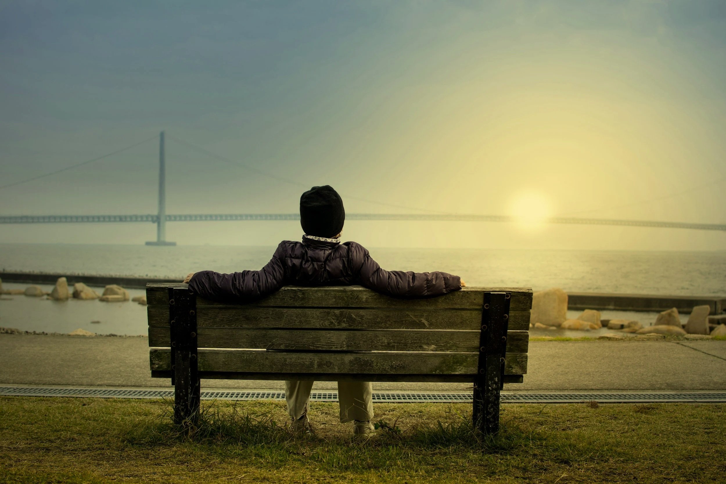 Person sitting in relaxed posture on a bench facing the water during sunset, with a bridge in the background evoking reflection and integration after psychedelic journey work.