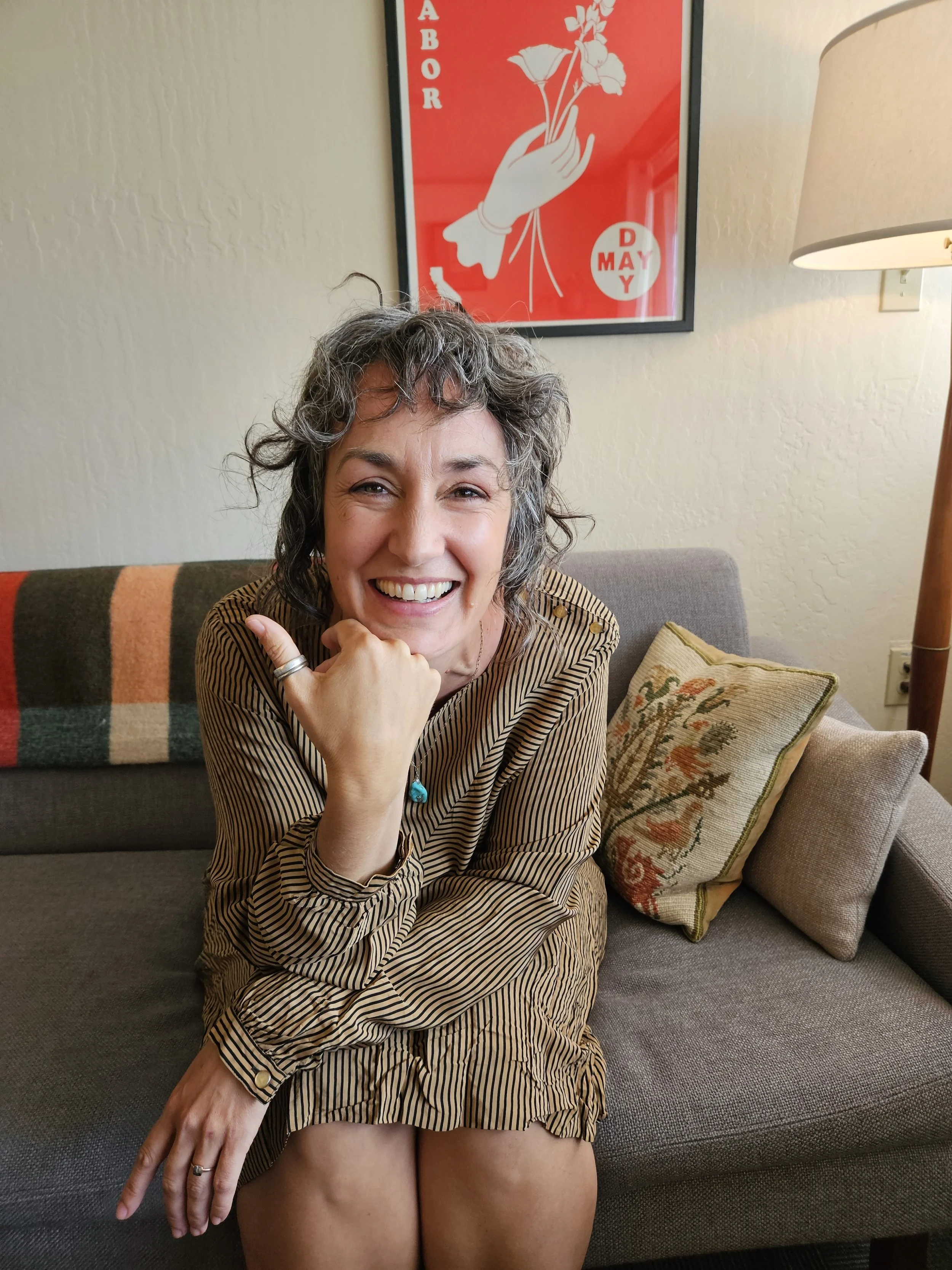 Carly Rose Bretl, LMFT, woman and therapist with curly gray hair smiling and sitting on a gray sofa in therapy office with a colorful blanket, decorative pillows, and a framed poster in the background.