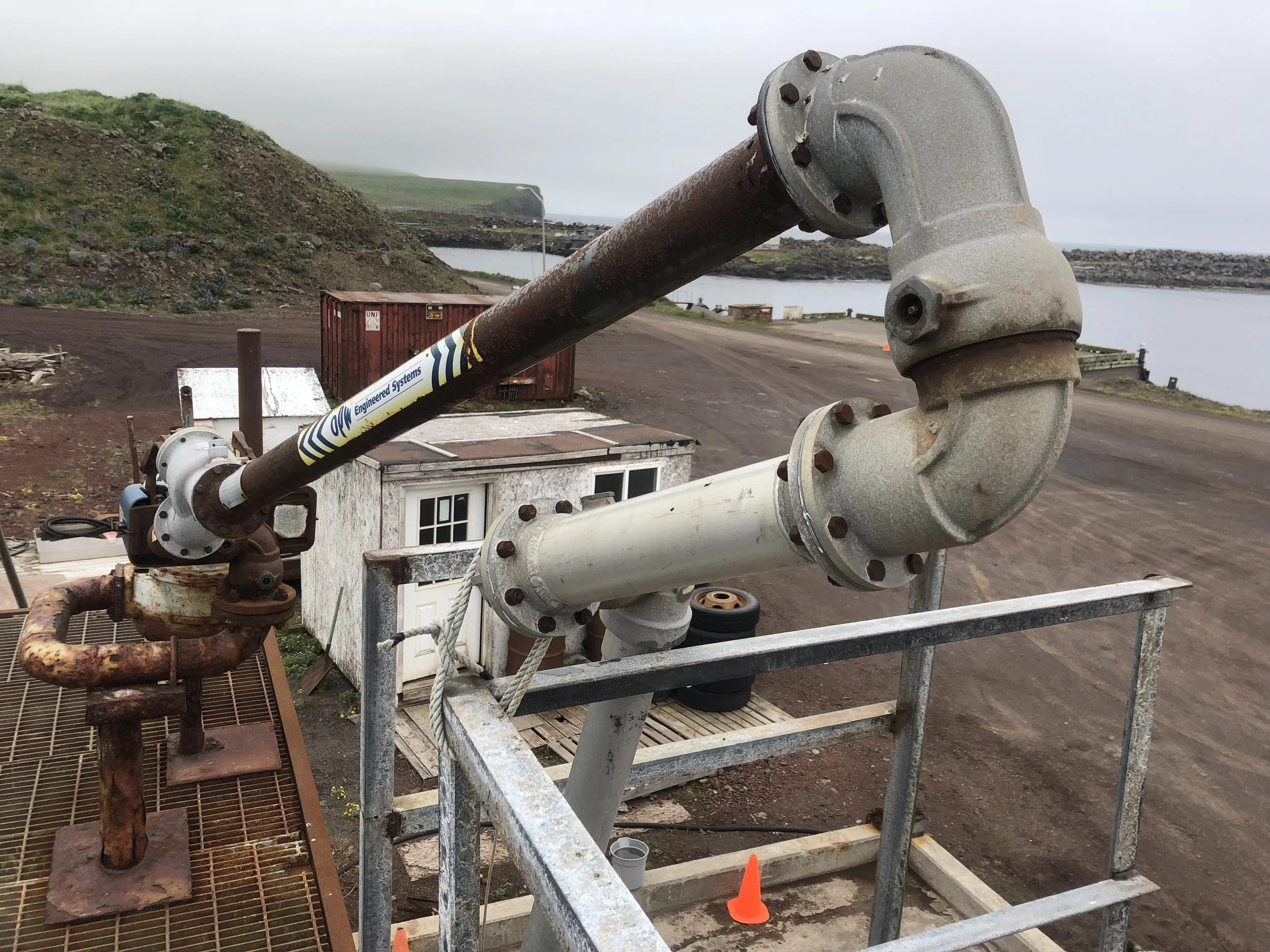 Large industrial pipe and valve system outdoors near a body of water with hills in the background.