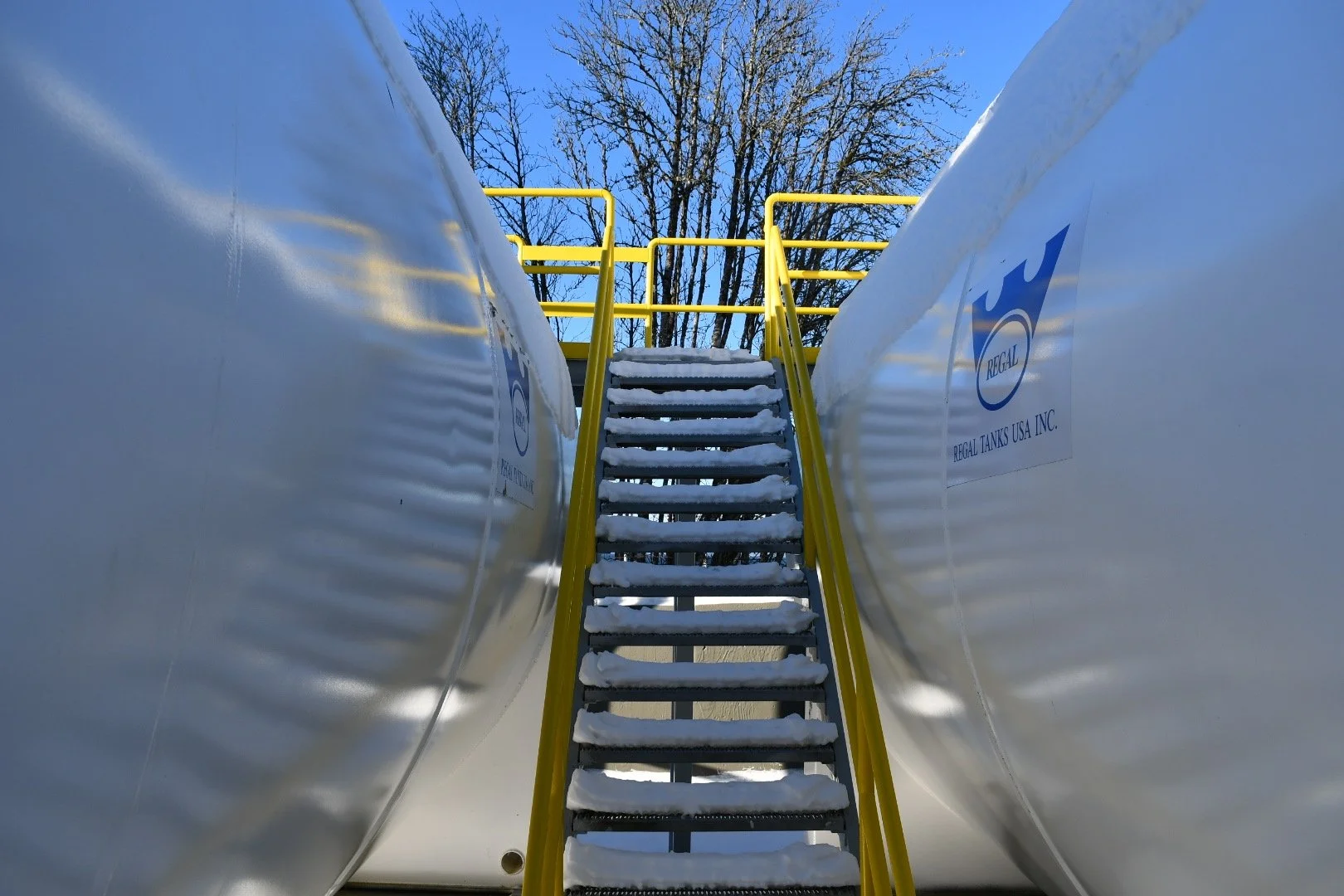 Looking up at a stainless steel storage tank with a ladder and yellow handrails in a snowy outdoor environment, with leafless trees in the background and a clear blue sky.