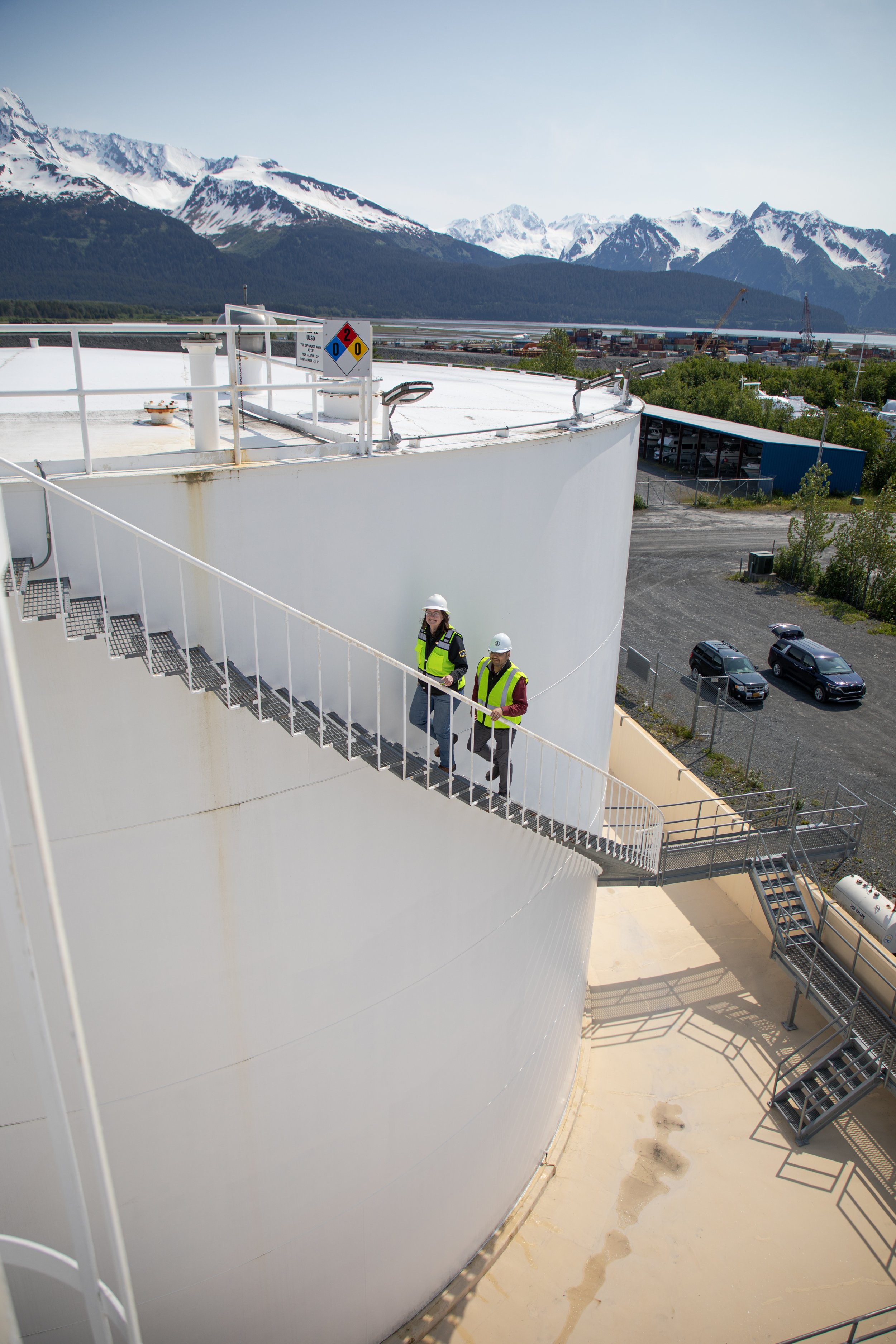 Two environmental compliance consultants descending a staircase on a large aboveground fuel storage tank with Alaska mountains in the background