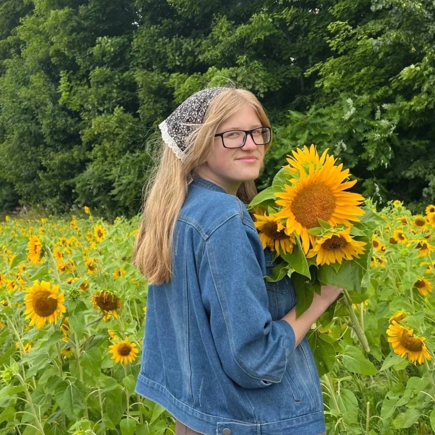 A woman with long blonde hair, glasses, and a black and white headscarf, standing in a sunflower field, holding a bouquet of sunflowers, with green trees in the background.