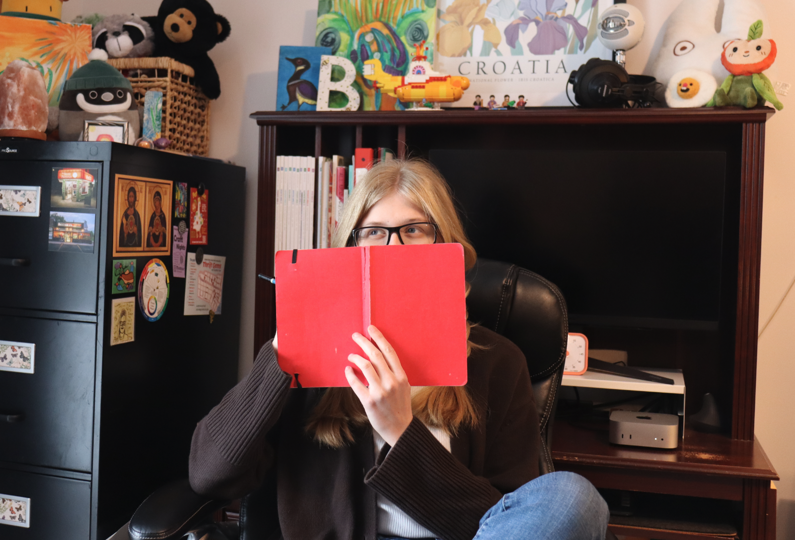 A woman with glasses holding a red tablet, sitting in a chair in a room with bookshelves, plush toys, and colorful artwork in the background.