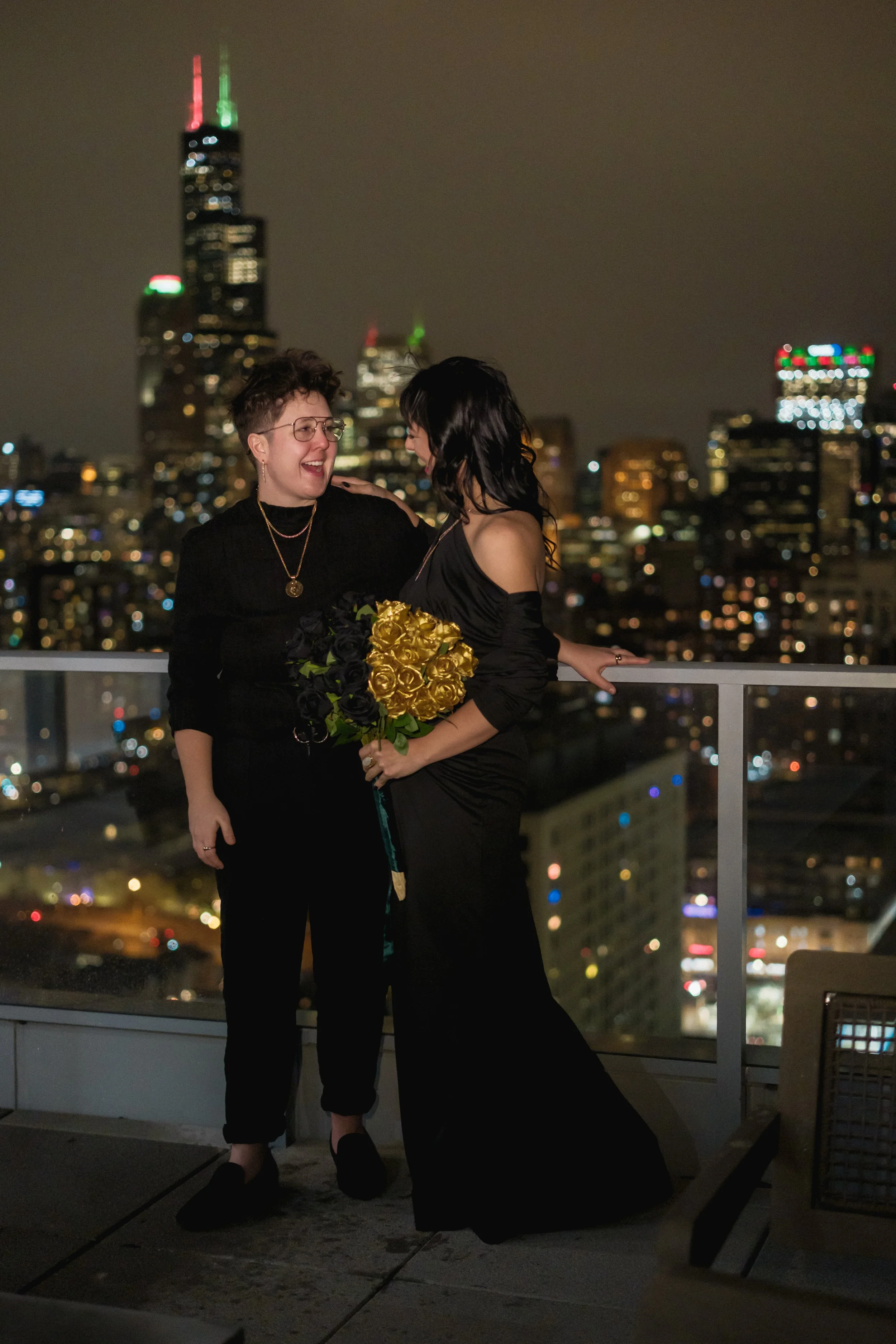 Two women in black dresses celebrating on a city rooftop at night, holding a bouquet of golden and black roses, with illuminated skyscrapers in the background.