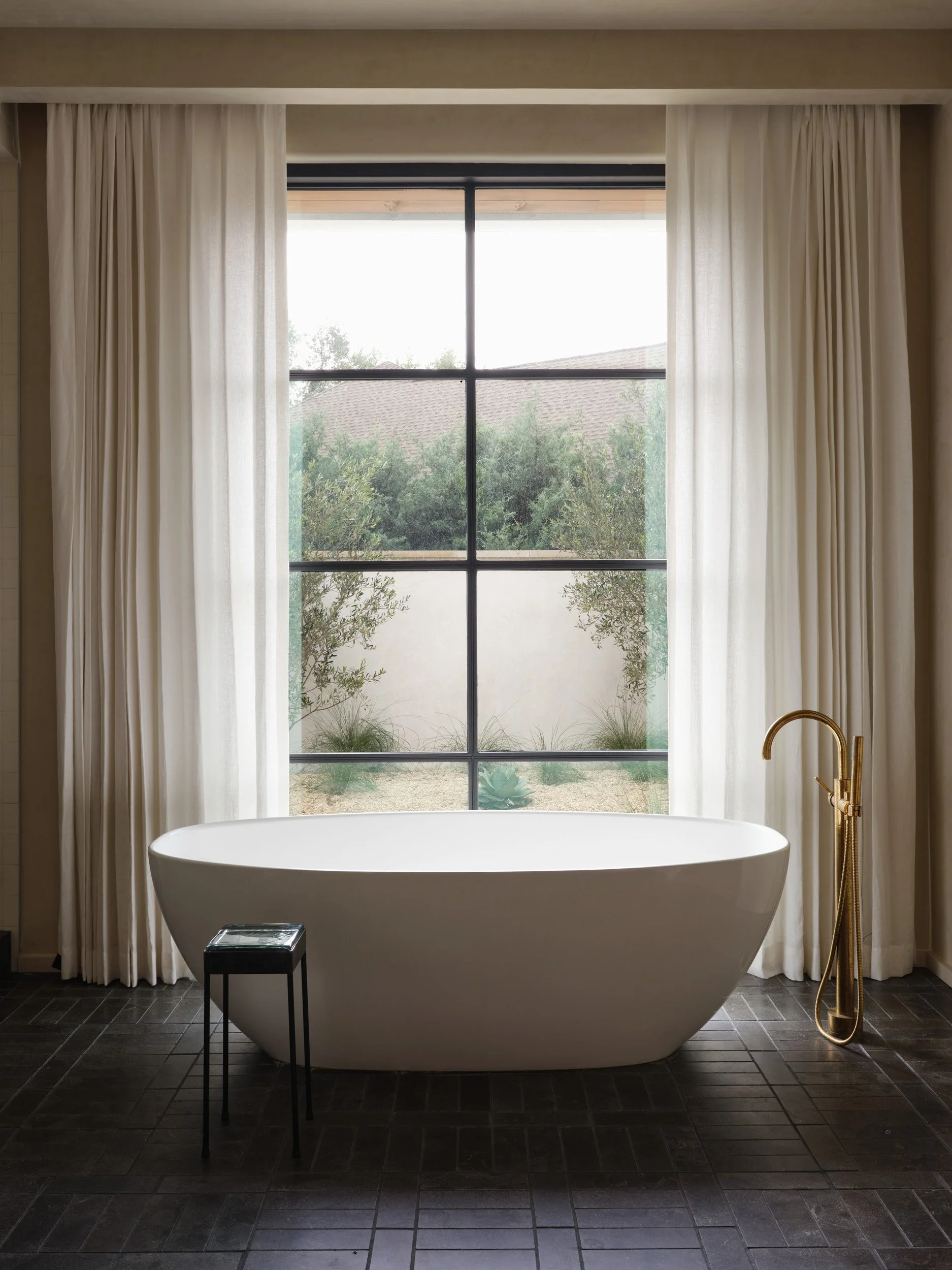 A modern bathroom with a freestanding white bathtub in front of a large window with sheer white curtains; a small black side table and a gold bathtub faucet are also visible.
