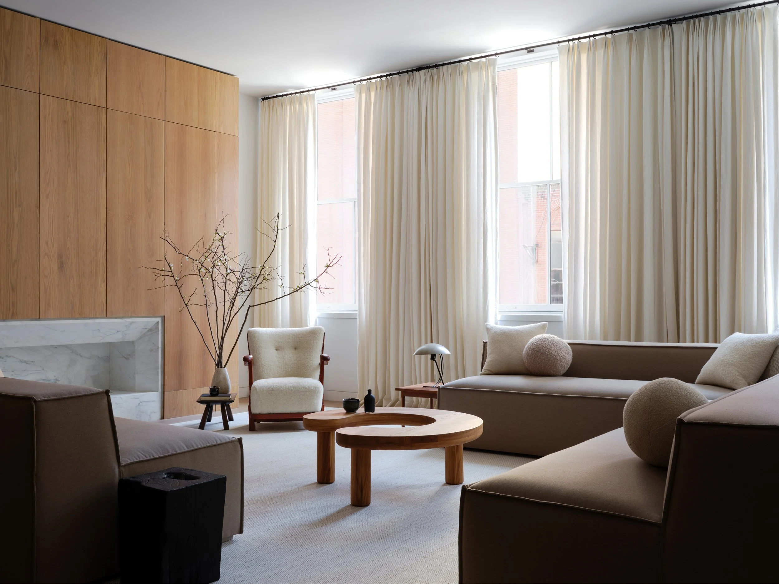 Modern living room with beige sofas, a wooden coffee table, a white armchair, and large windows with cream curtains.