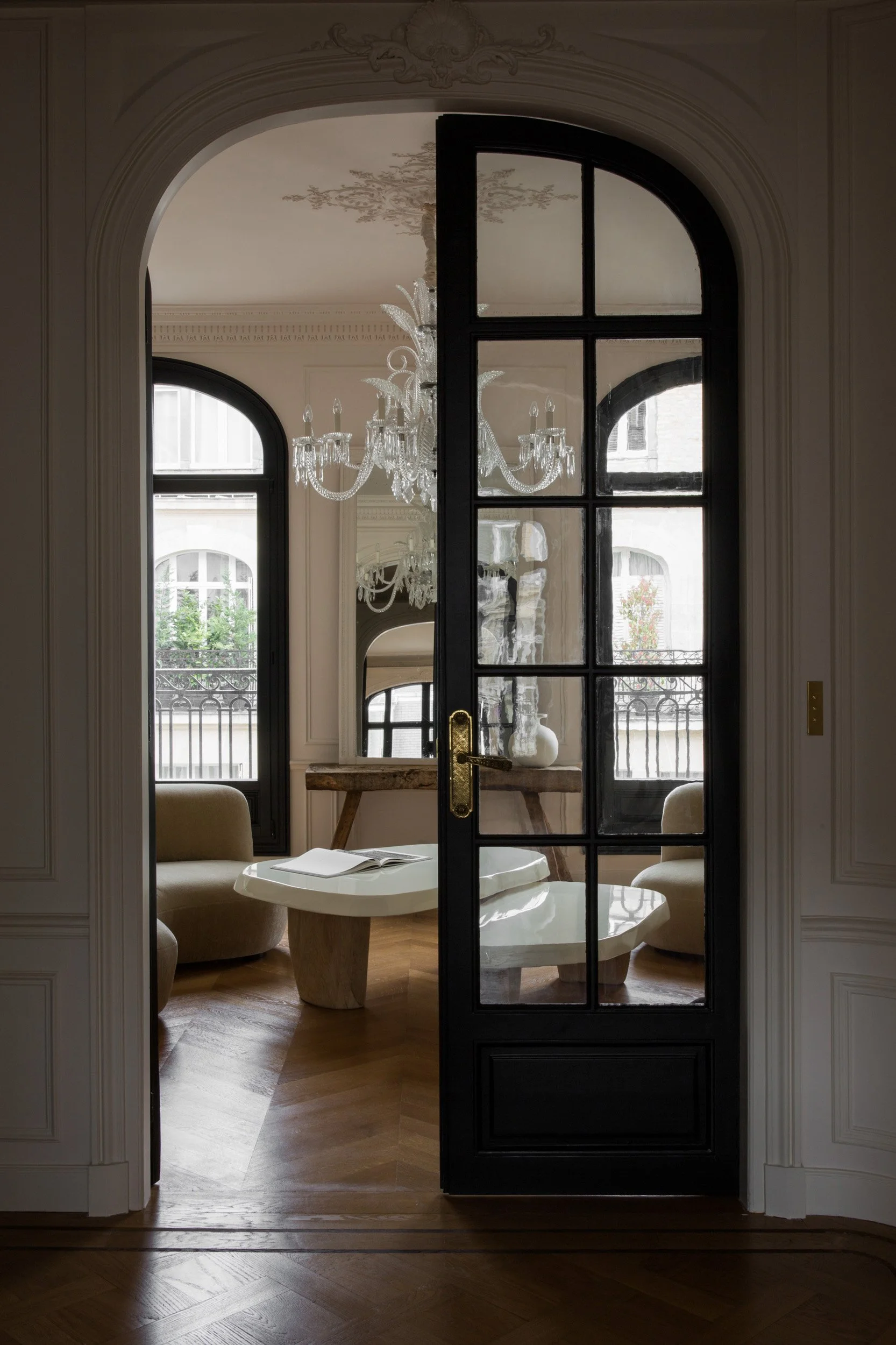 A view through a black glass-paneled door into a luxurious sitting area with a chandelier, a white marble table, cushioned chairs, and large arched windows.